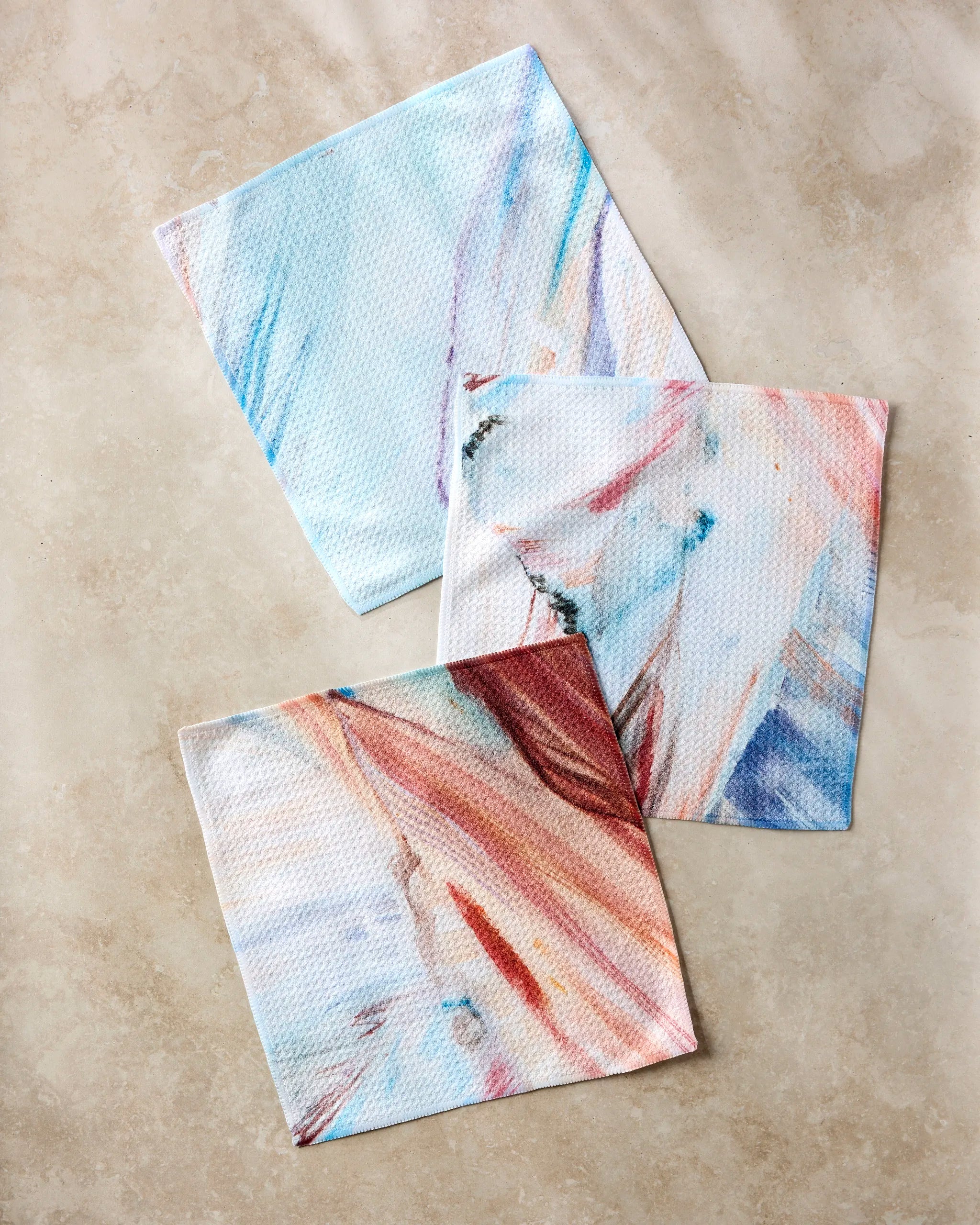 Three super-absorbent cloths from the "Golden Hour in the Rockies" dishcloth set by Kelly Peters, featuring abstract pastel patterns in blue, pink, and red, are displayed on a beige marble-like surface. Made from recycled materials.