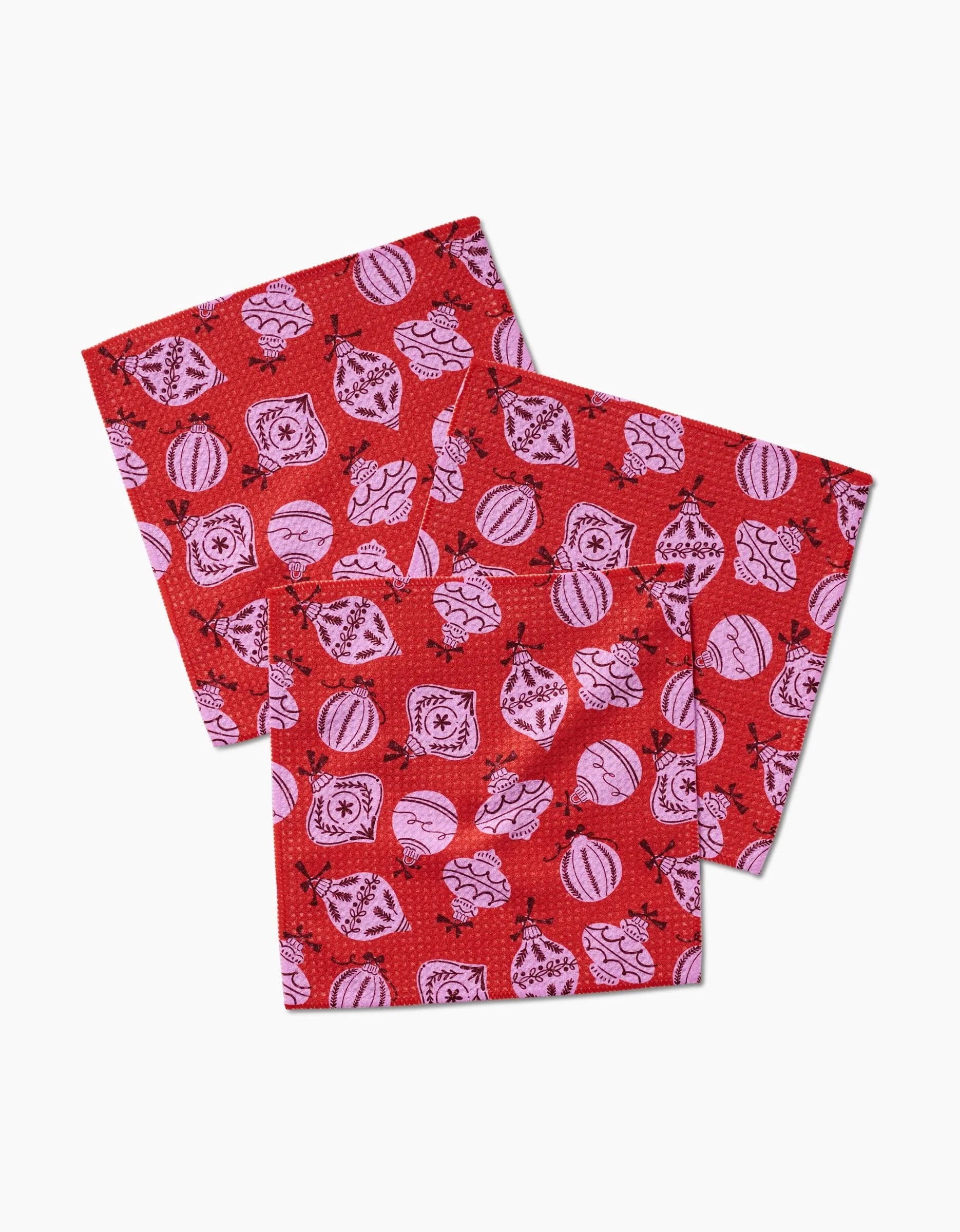 Three Grams Ornaments napkins by Charlie Rowan Designs, featuring red and pink Geoweave fabric with Christmas ornaments and festive patterns, are stacked slightly askew on a white background.