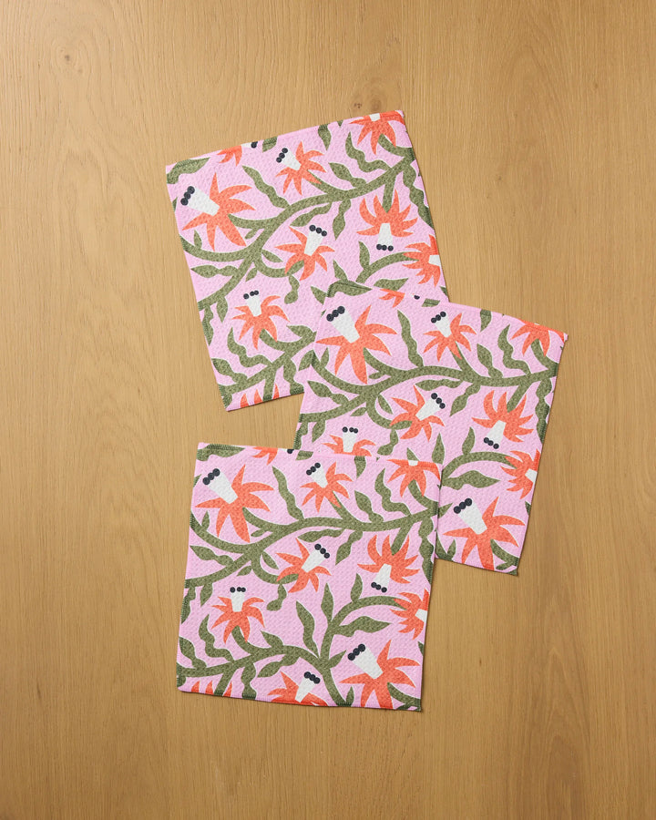 The Claire Iglesias "Graphic Flowers" set features three recycled cloth napkins with a pink background, red-orange flowers, and green leaves—an elegant addition to your kitchen.