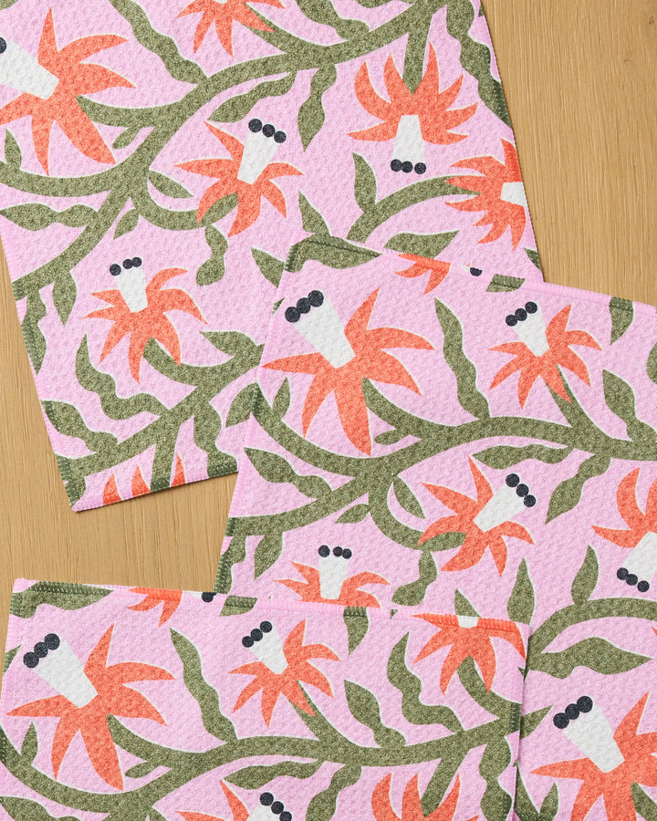Four absorbent "Graphic Flowers" kitchen towels by Claire Iglesias, featuring a pink pattern with stylized orange flowers, green stems and leaves, plus black dots, arranged overlapping on a light wooden surface.