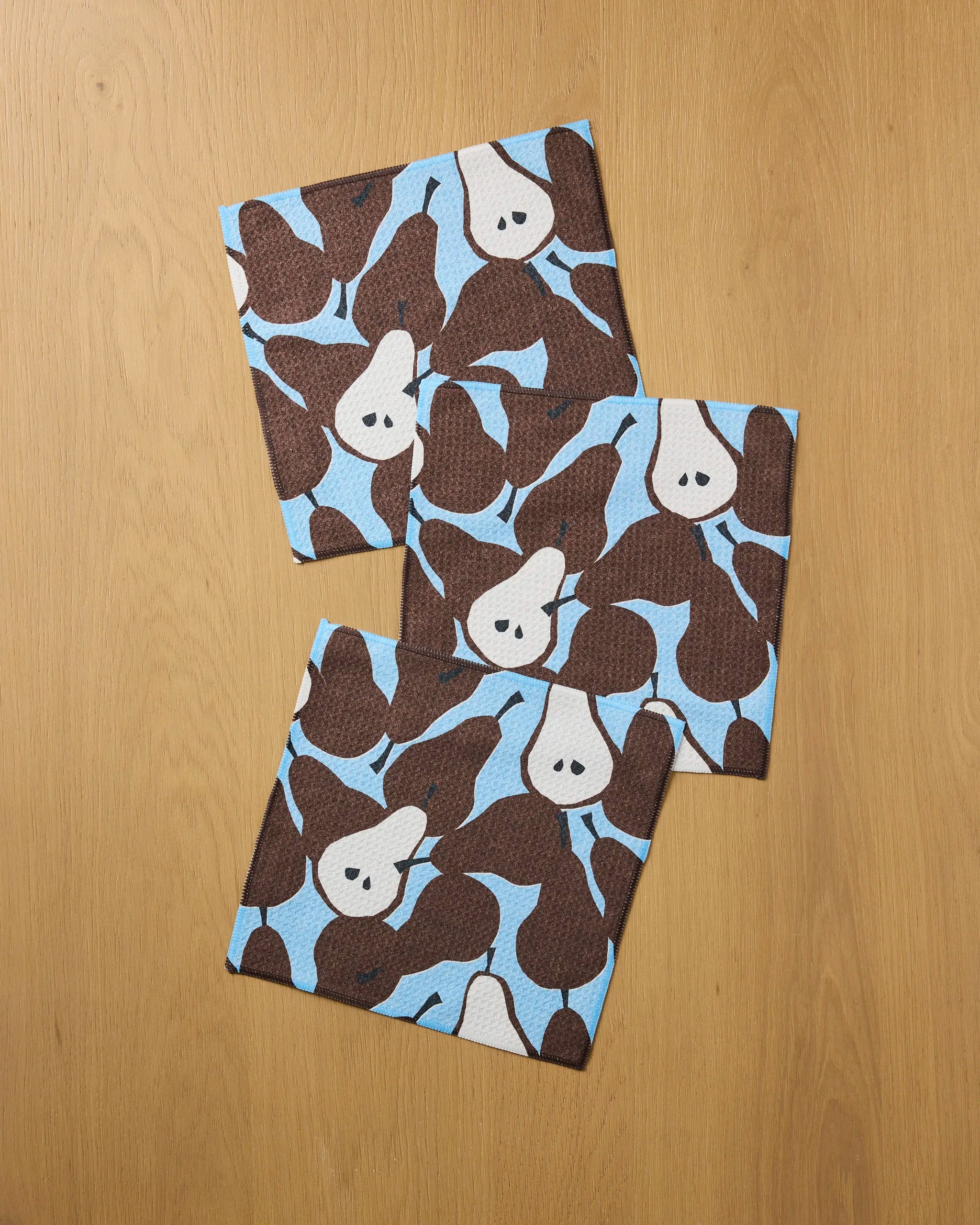 Three Graphic Pears napkins by Claire Iglesias, made from recycled materials, display a brown and white pear pattern on light blue and are arranged on a wooden surface.