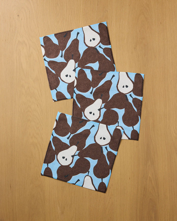 Three Graphic Pears napkins by Claire Iglesias, made from recycled materials, display a brown and white pear pattern on light blue and are arranged on a wooden surface.