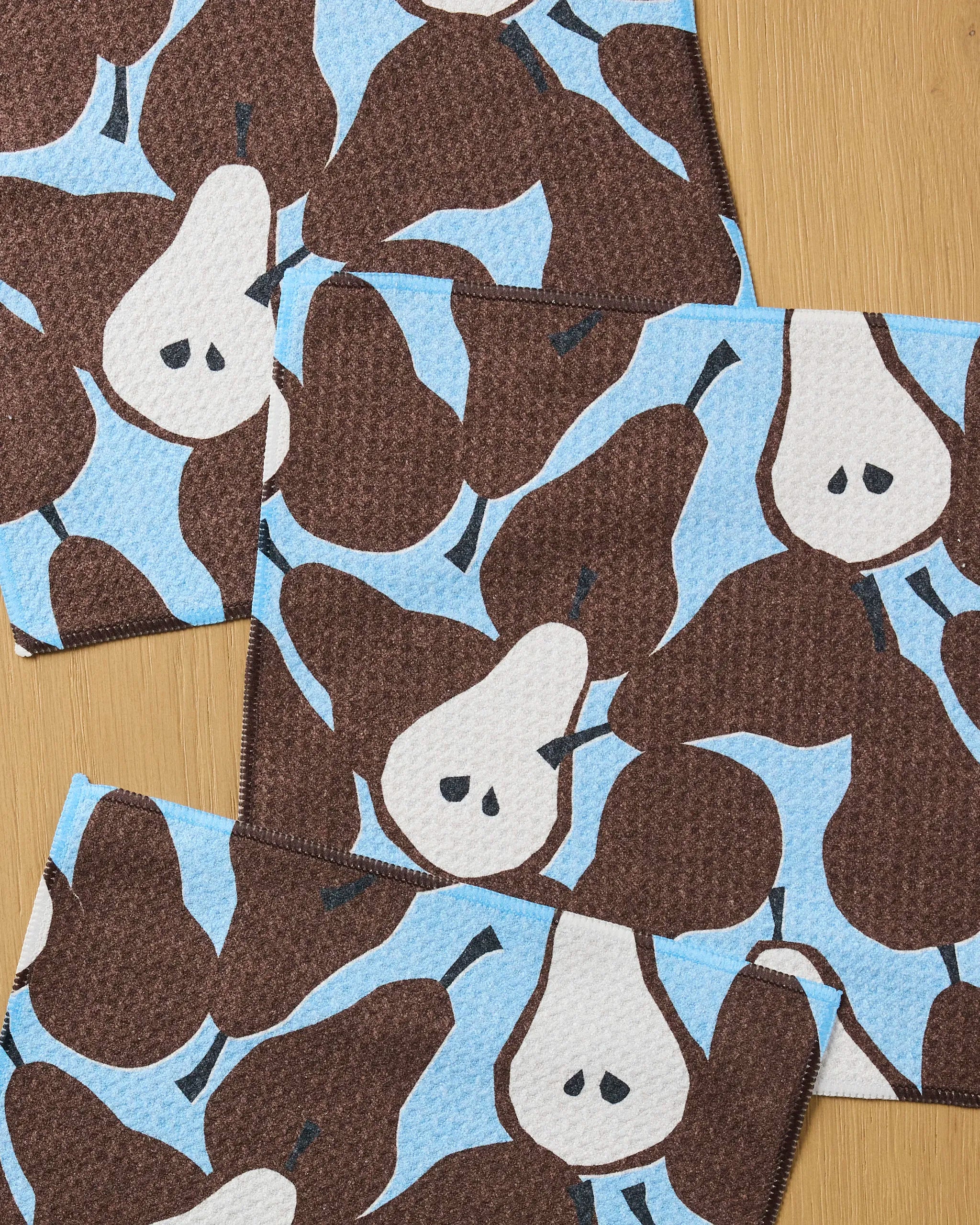 The Claire Iglesias Graphic Pears set includes four super-absorbent dishcloths, each featuring a repeating brown pear pattern on light blue, displayed on a wooden surface.