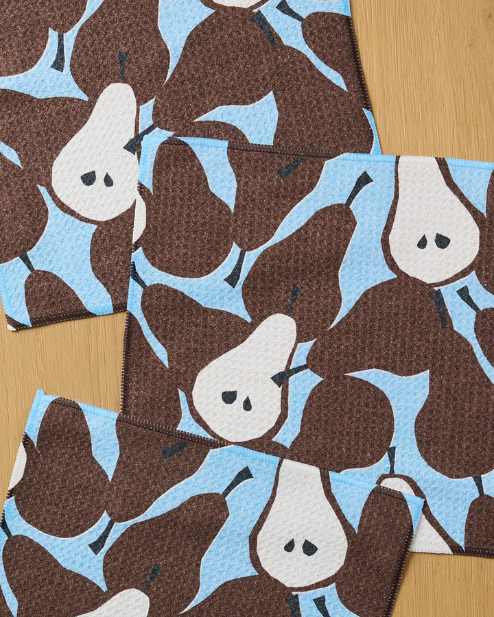 The Claire Iglesias Graphic Pears set includes four super-absorbent dishcloths, each featuring a repeating brown pear pattern on light blue, displayed on a wooden surface.