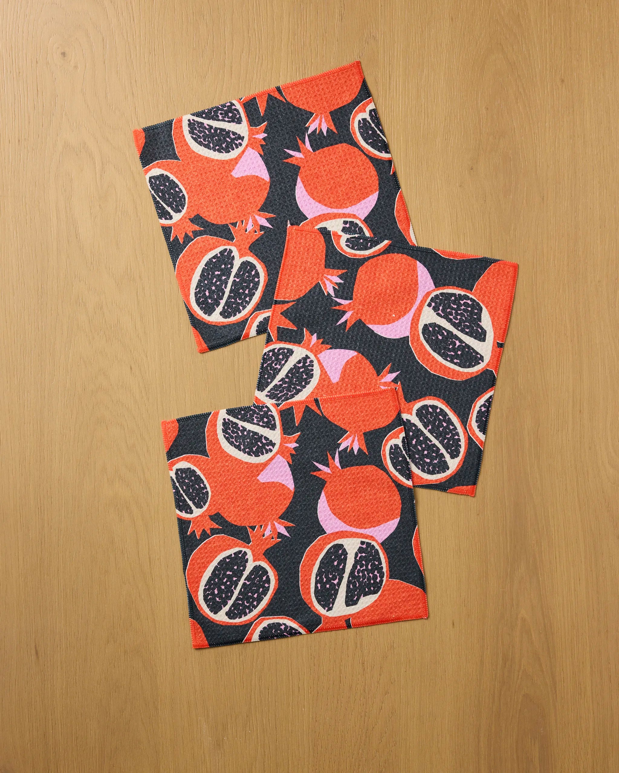 Three folded “Graphic Pomegranates” napkins by Claire Iglesias, featuring a bold red, pink, and black pattern, are arranged on a light wood surface—perfect for adding vibrant color to your dishcloth set.