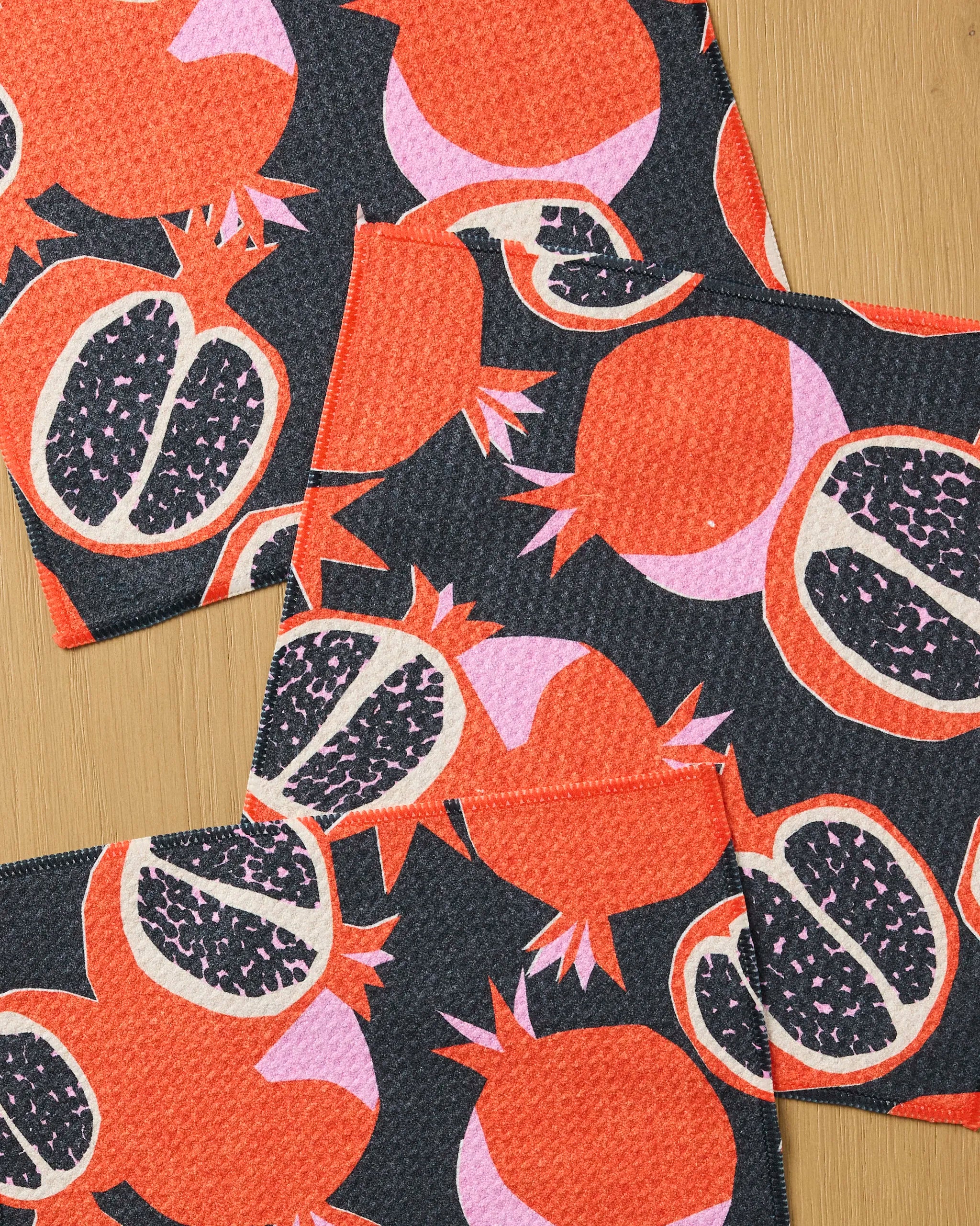 Three “Graphic Pomegranates” cloth napkins by Claire Iglesias, featuring a bold red, pink, and black pattern, are displayed on wood. This vibrant set combines playful style with super absorbency for functional, everyday use.