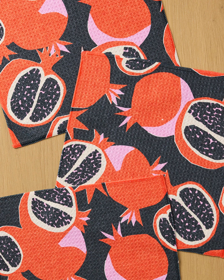 Three “Graphic Pomegranates” cloth napkins by Claire Iglesias, featuring a bold red, pink, and black pattern, are displayed on wood. This vibrant set combines playful style with super absorbency for functional, everyday use.