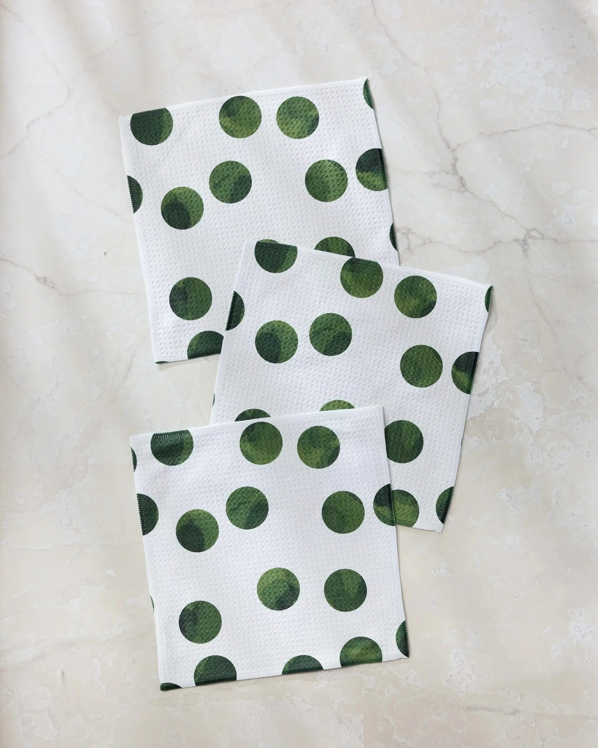 Three "Grass Polka" napkins by Geometry, featuring large green polka dots on white and made from recycled materials, are shown slightly overlapping on a light marble surface.