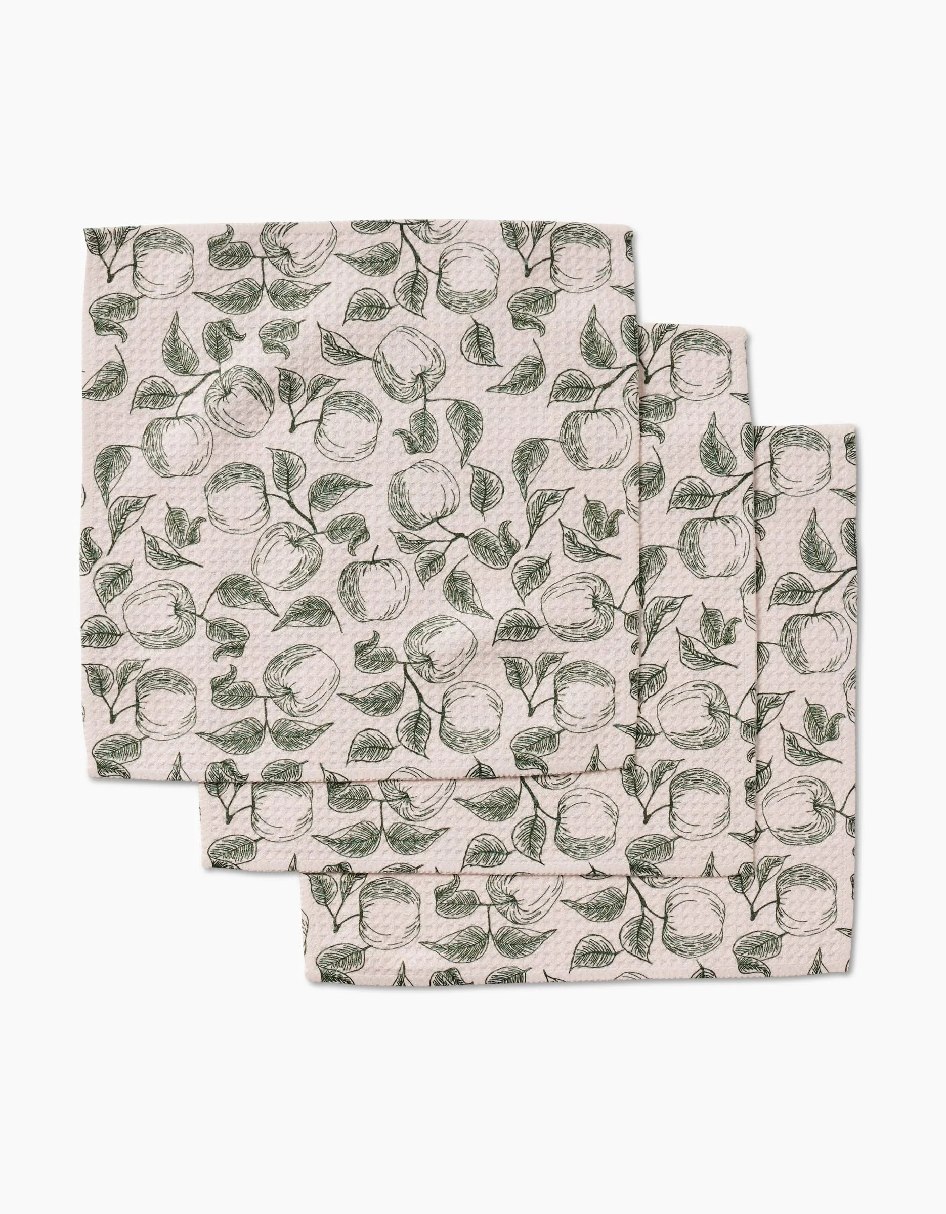 Three folded Gravenstein Apple cloth napkins by Ash Cascade, made from Geoweave fabric, feature a repeating hand-drawn apple and leaf pattern in green on a light beige background, arranged with slight overlap.