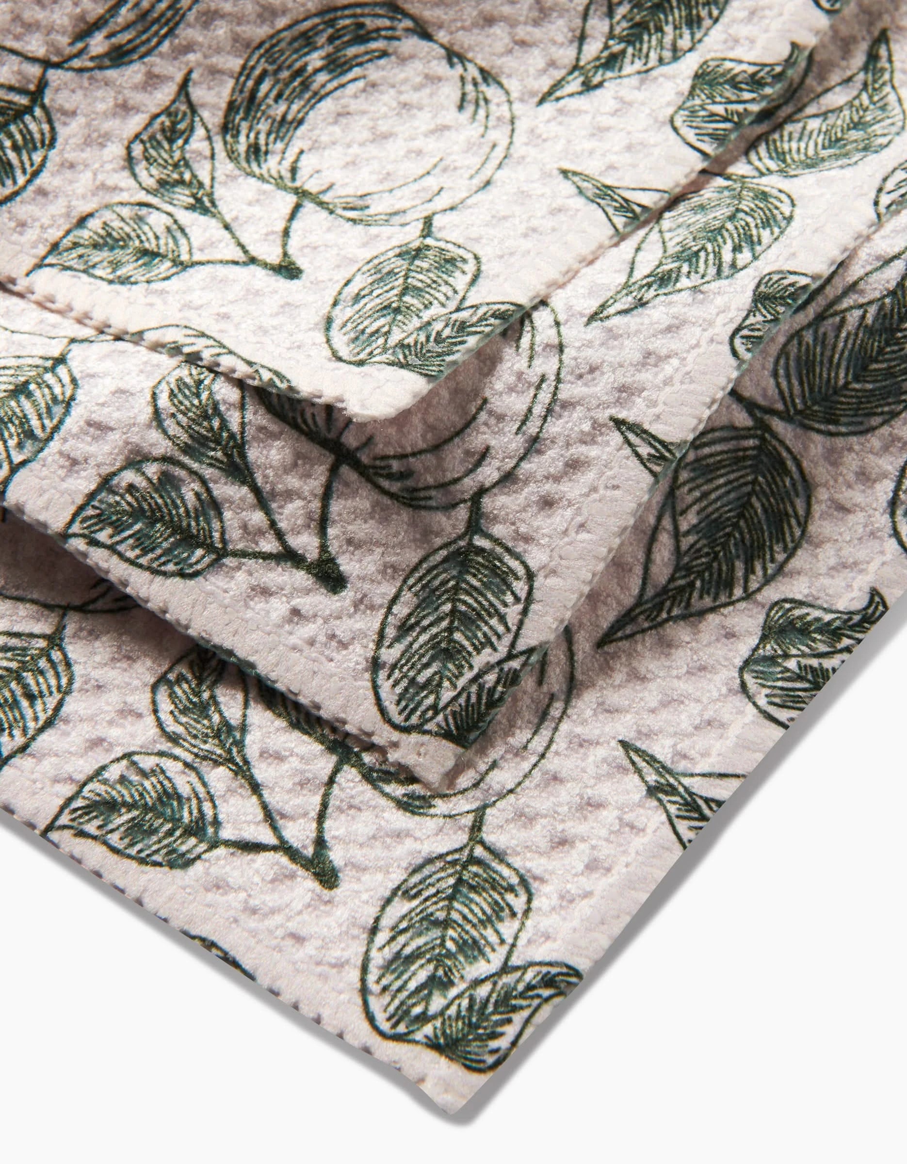 A close-up of folded, textured Gravenstein Apple dishcloth by Ash Cascade, featuring a green leaf pattern on light beige recycled Geoweave fabric. The visible edges highlight the repeating design and soft, eco-friendly material.