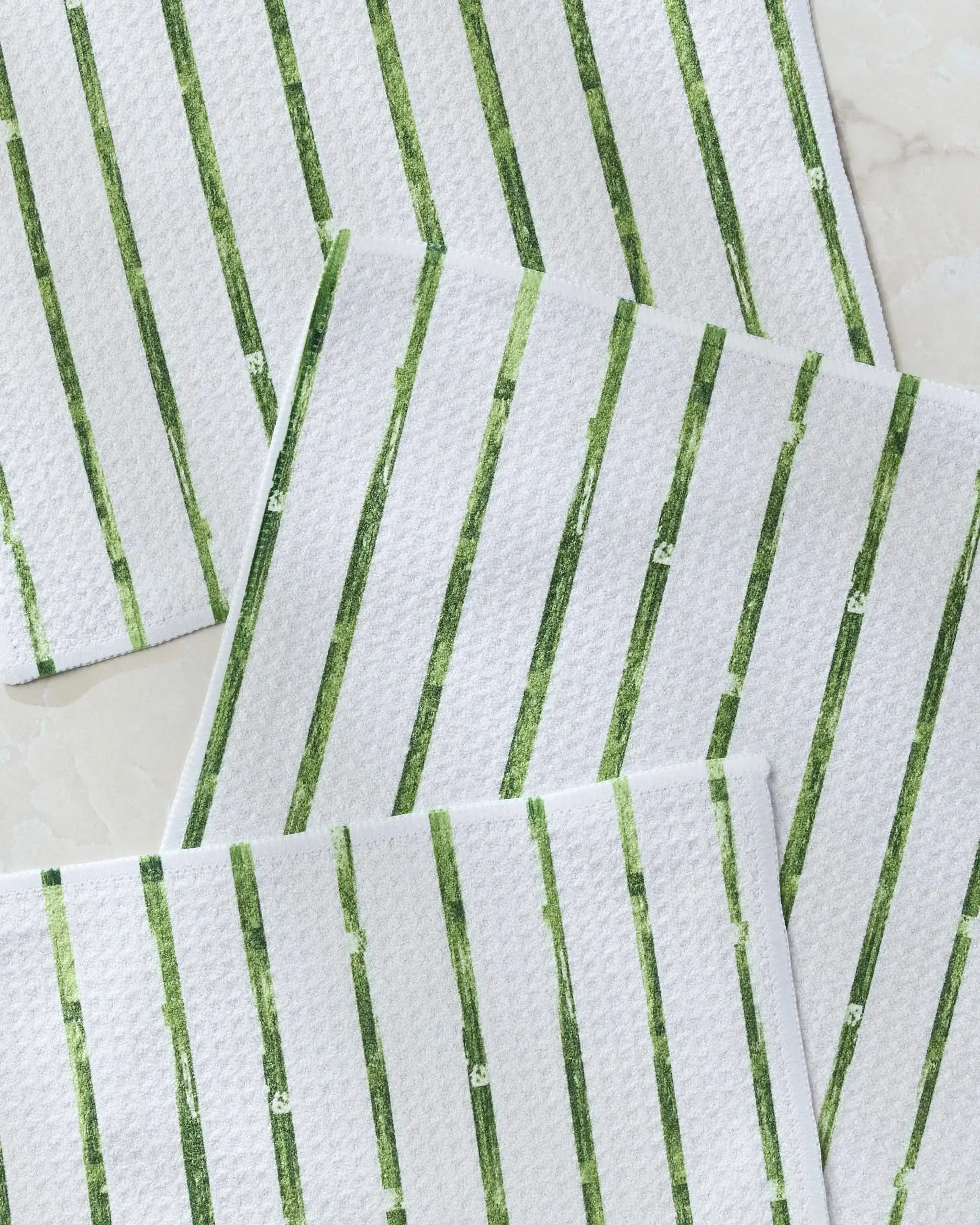 Four Greenhouse Stripe kitchen towels by Geometry, featuring green vertical stripes on white, are artfully overlapped on a light surface, showcasing a stylish and practical classic dishcloth set.