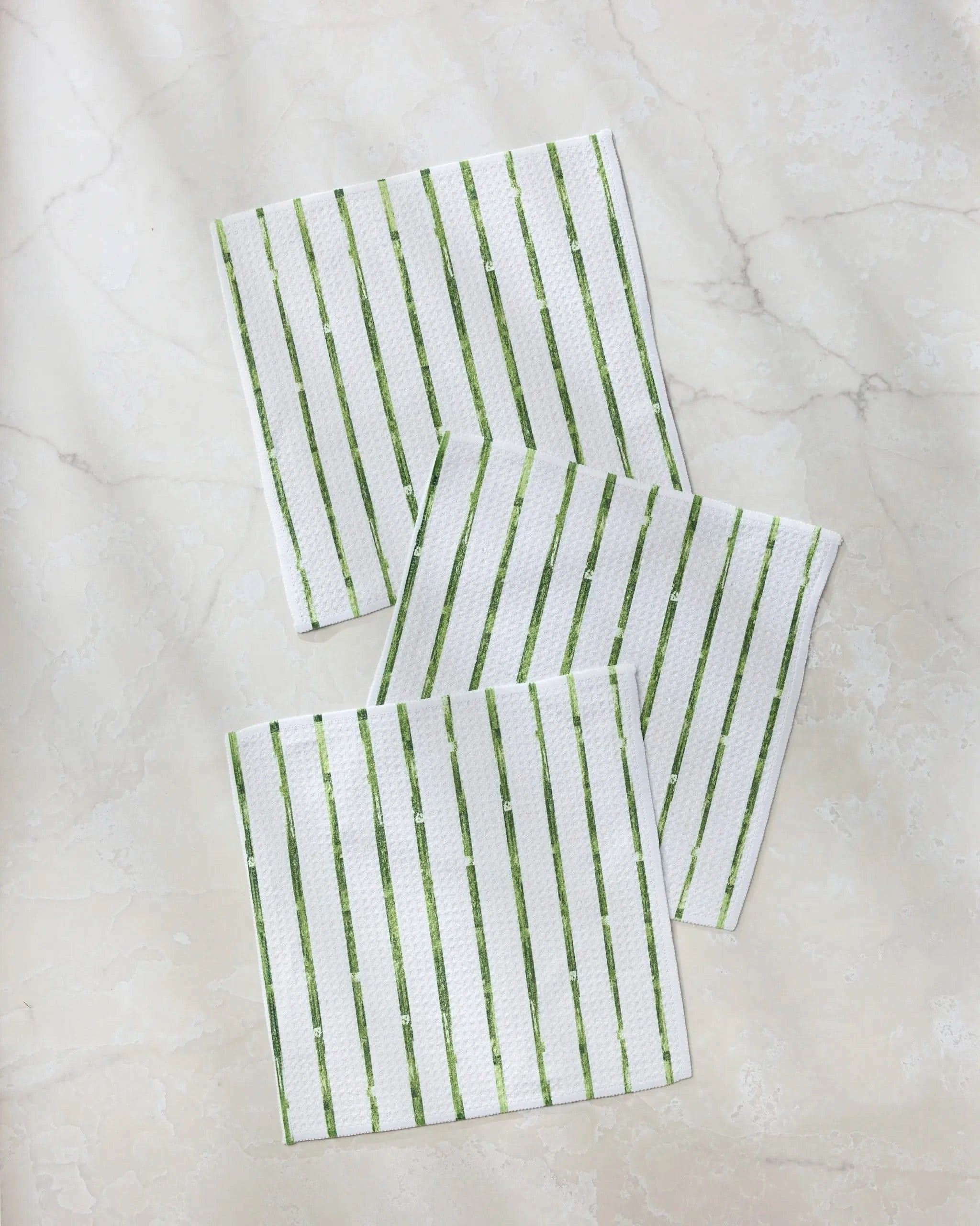 Three white Geometry dishtowels from the Greenhouse Stripe collection, featuring green vertical stripes, are arranged on a light beige, marble-patterned surface.