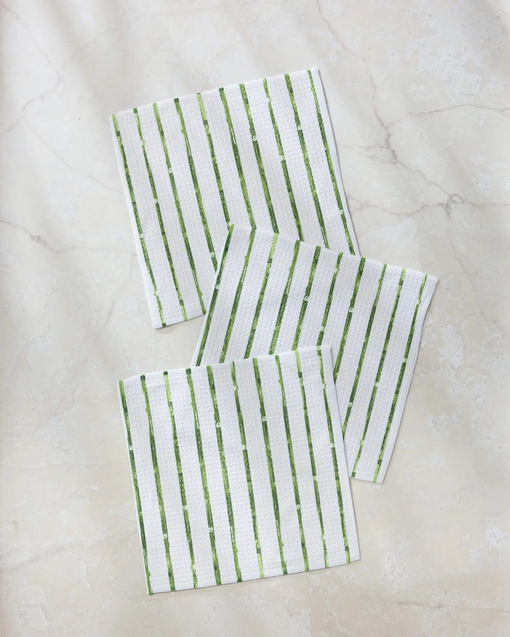 Three white Geometry dishtowels from the Greenhouse Stripe collection, featuring green vertical stripes, are arranged on a light beige, marble-patterned surface.