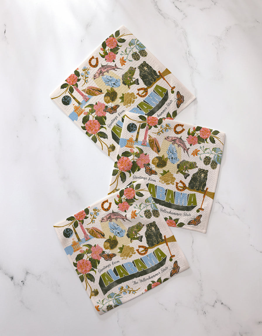 The Geometry "Greetings From Alabama" set includes three recycled square dishcloths featuring colorful Alabama-themed illustrations—flowers, birds, and landmarks. Each displays “Alabama” on a crisp white background.