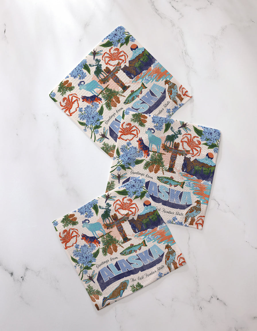 Three square "Greetings From Alaska" napkins by Geometry, featuring colorful Alaska-themed illustrations and the word ALASKA, are arranged on white marble—perfect as cheerful recycled dishcloths or a kitchen set.
