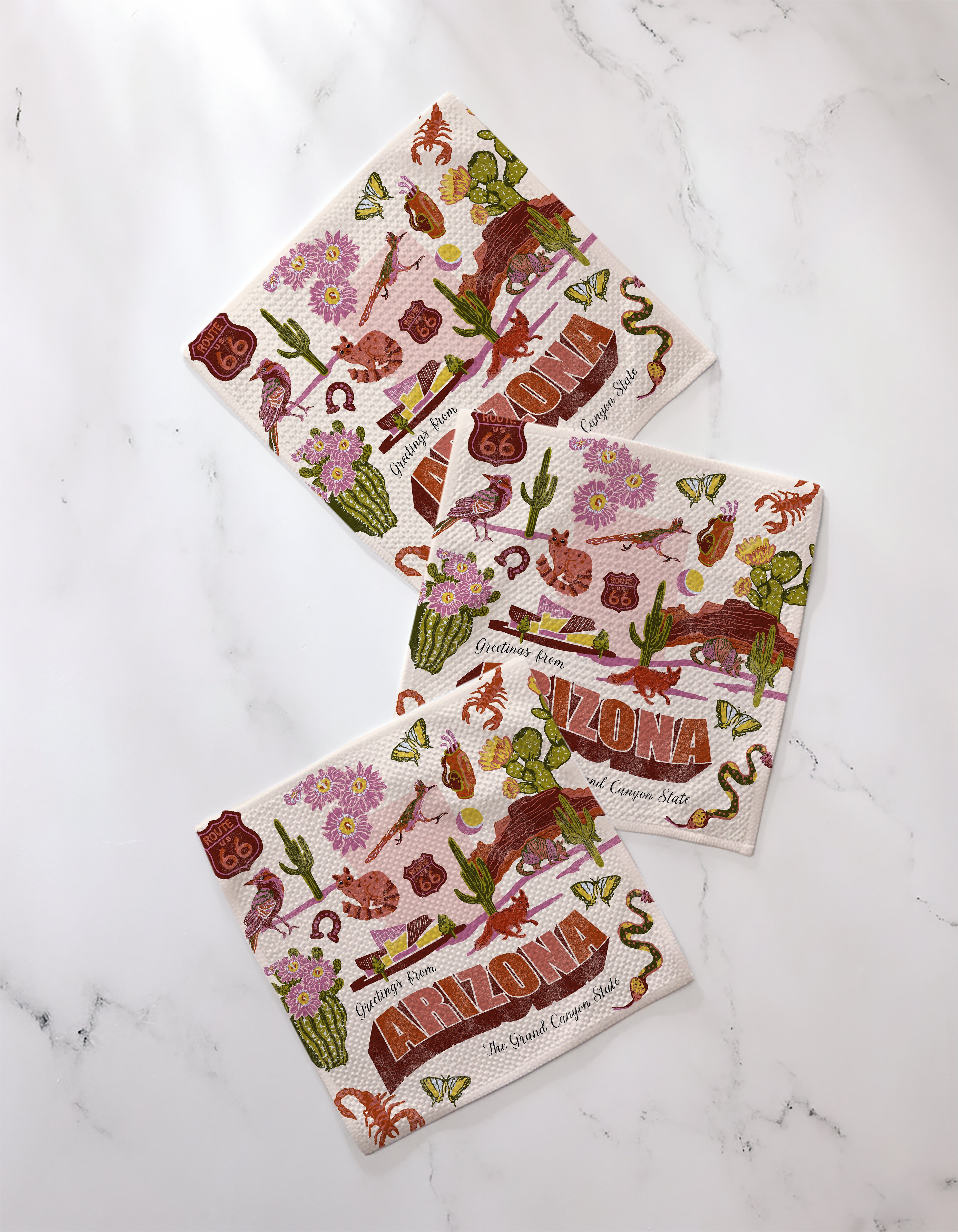 Three "Greetings From Arizona" paper napkins by Geometry, featuring bold ARIZONA lettering with cacti, flowers, snakes, birds, and Route 66 signs—perfect for pairing with your favorite absorbent dishcloths.