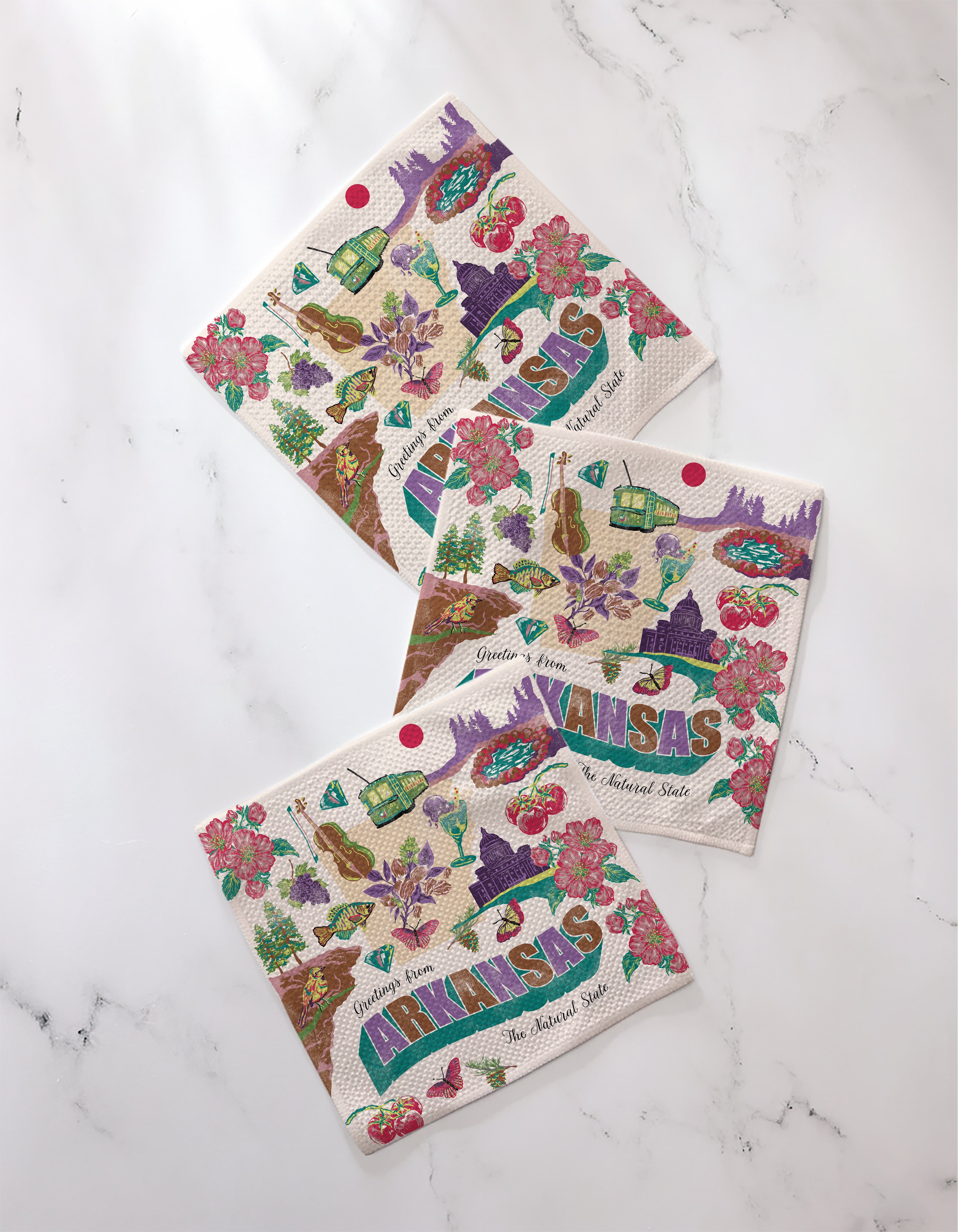 Three vibrant "Greetings From Arkansas" napkins by Geometry, crafted from recycled materials and adorned with playful state icons, flowers, and landmarks, are displayed on a white marble-textured surface.