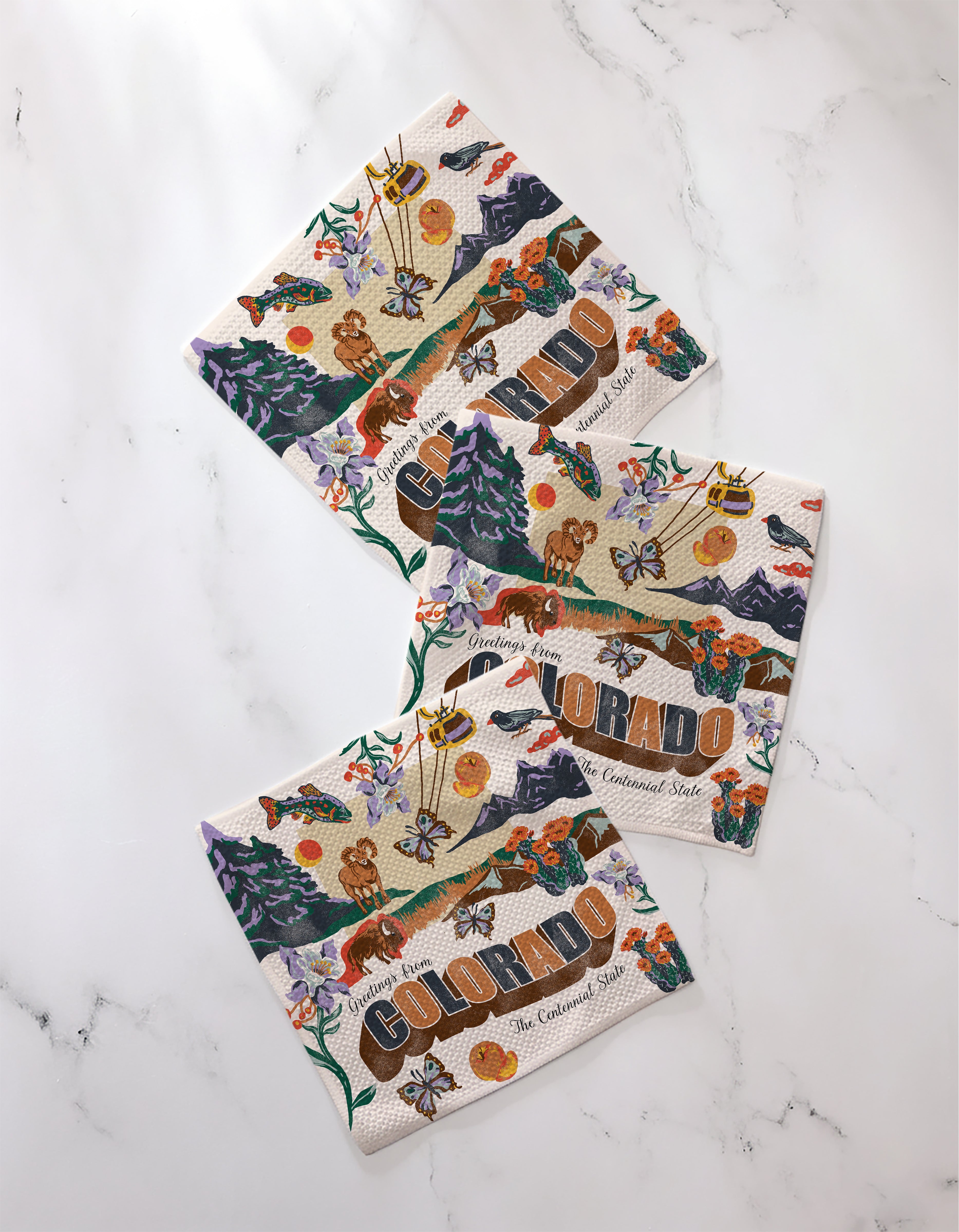 Four “Greetings From Colorado” decorative napkins by Geometry, made from recycled materials and featuring vibrant mountain, flower, wildlife, and state symbol illustrations, are displayed on a white marble surface.