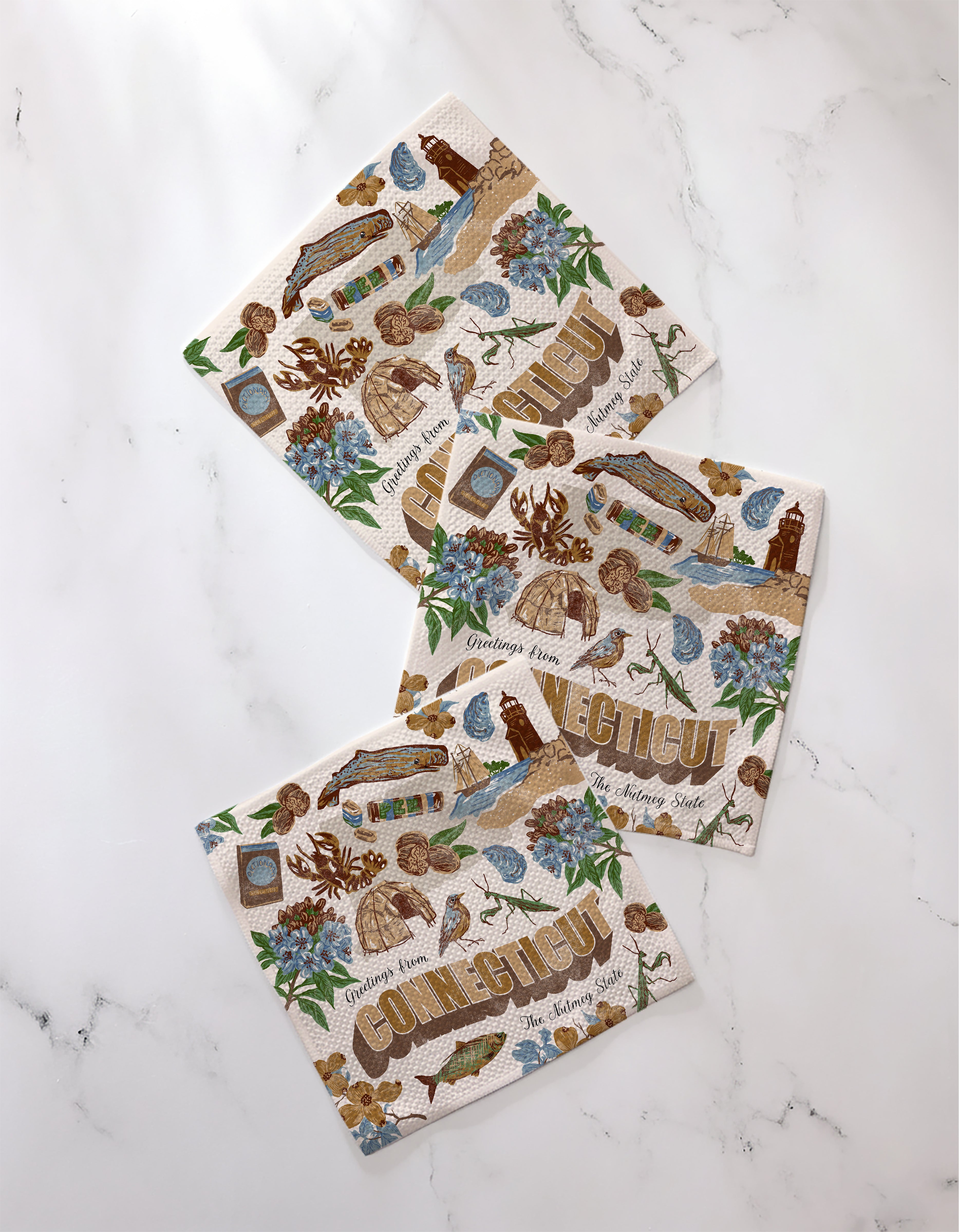 Three "Greetings From Connecticut" napkins by Geometry, made from recycled materials and decorated with illustrations and text inspired by Connecticut, are displayed on a white marble surface.
