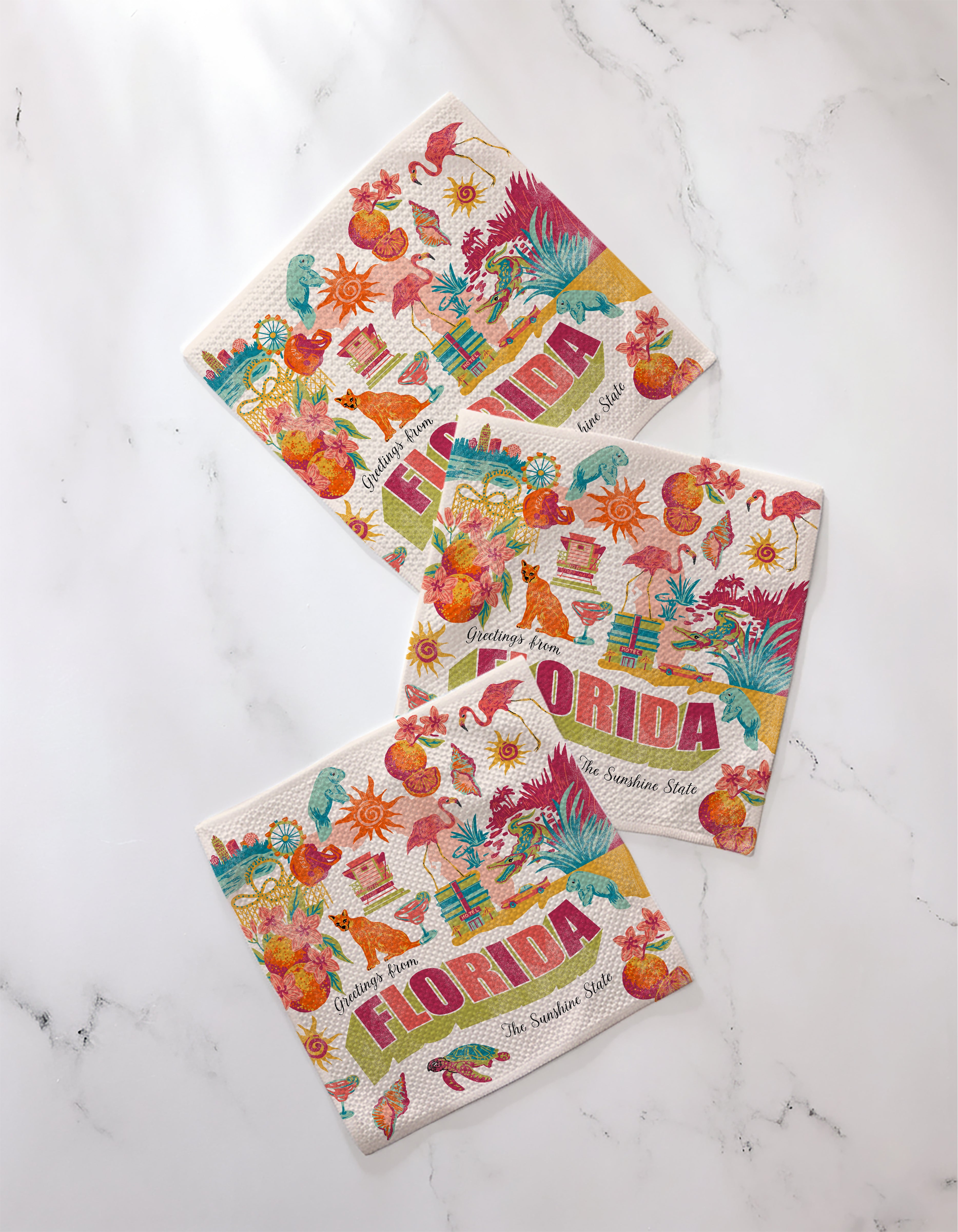The Geometry Greetings From Florida set features three colorful recycled dishcloths with tropical designs—flamingos, oranges, sunshine, palm trees, and hibiscus flowers—to brighten any kitchen. Displayed on a white marble surface.