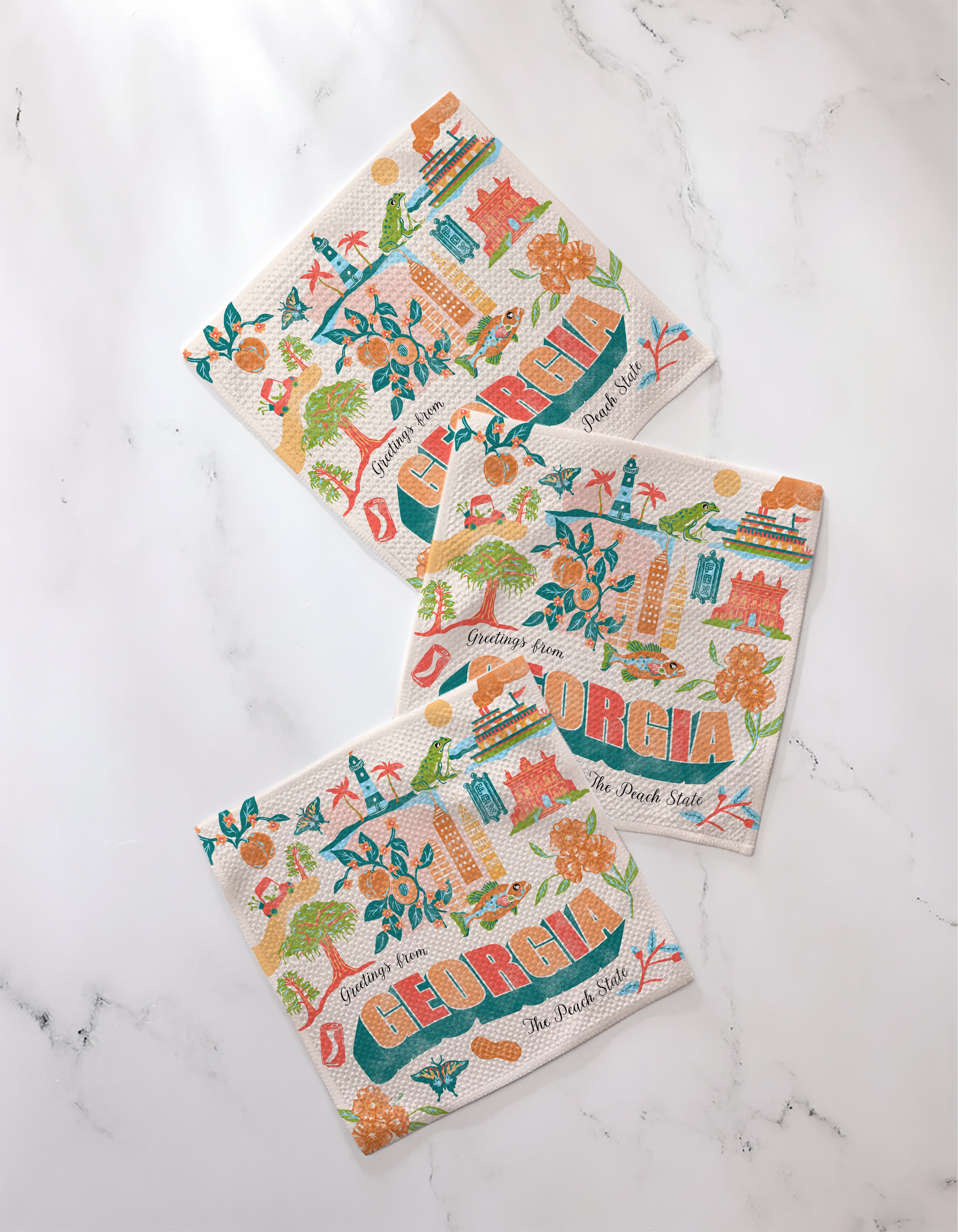 Three absorbent dishcloths from Geometry's "Greetings From Georgia" set, featuring colorful Georgia-inspired illustrations and “GEORGIA” in bold letters, are displayed on a white marble surface.