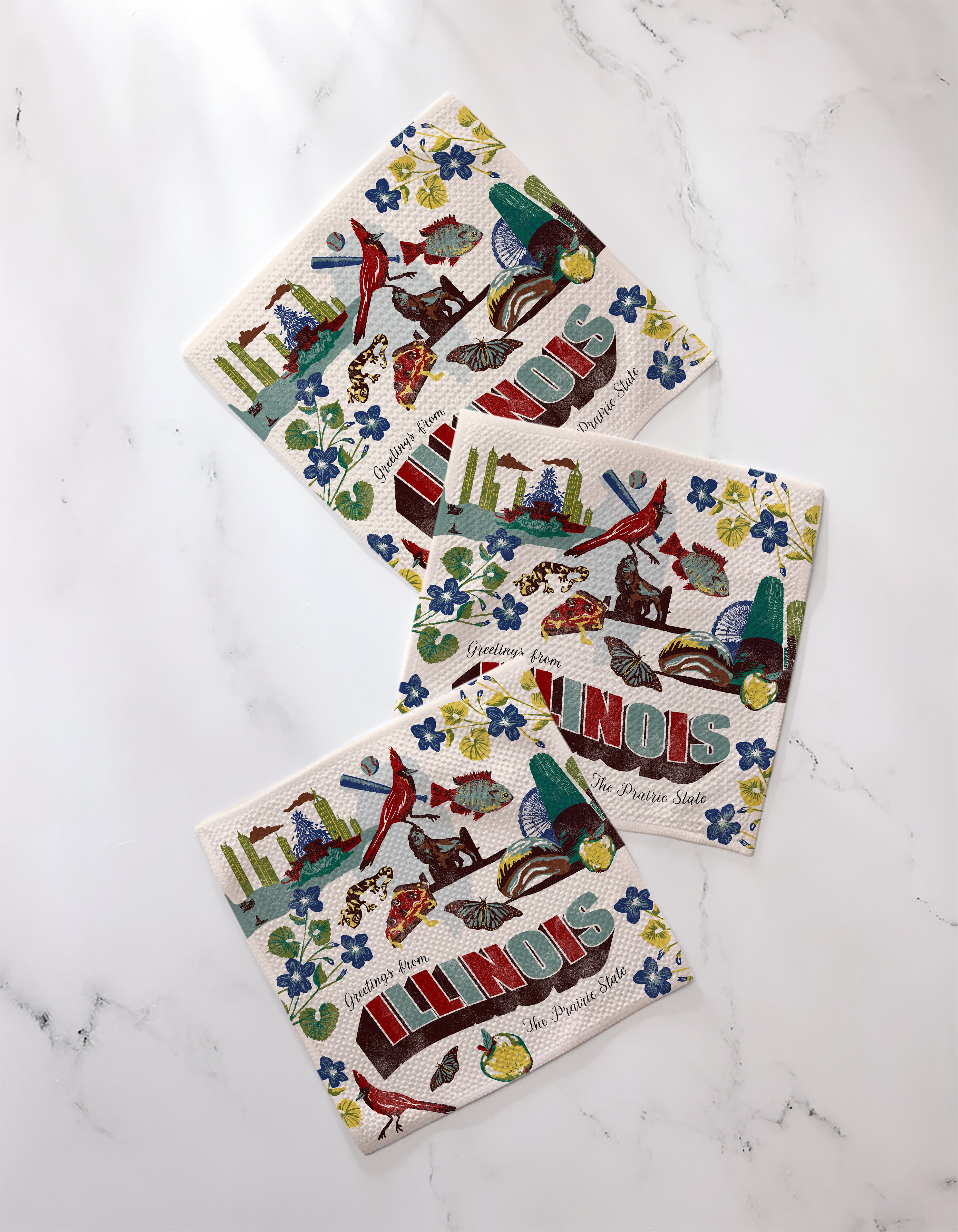 The Geometry Greetings From Illinois set features three illustrated, absorbent kitchen towels with ILLINOIS text, cardinals, Abraham Lincoln, flowers, and state symbols plus "Greetings from The Prairie State" on a marble surface.
