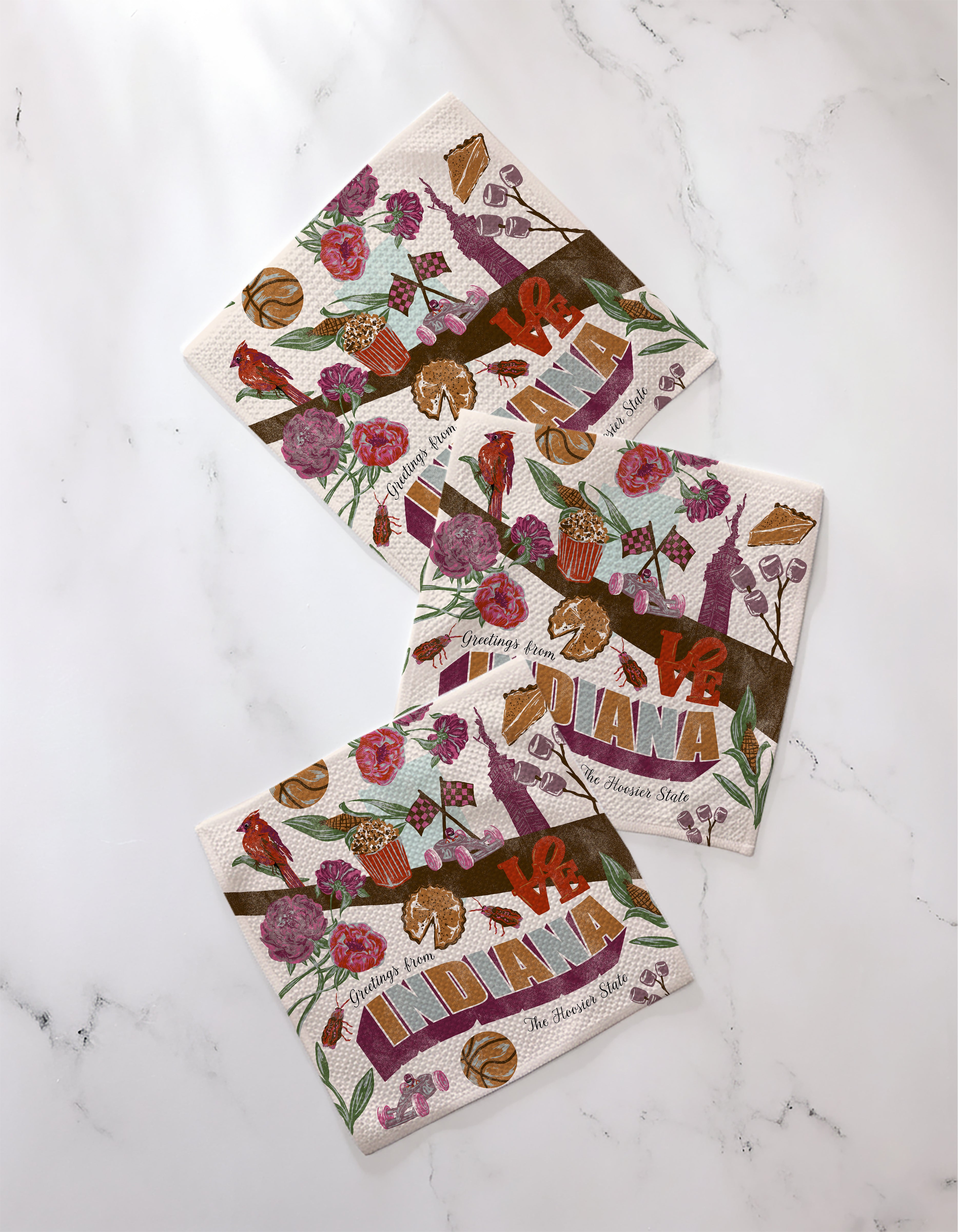 The Geometry "Greetings From Indiana" decorative napkins feature recycled materials, floral, basketball, and bird illustrations, plus state text—making a unique addition to any dishcloth set. Shown on a white marble surface.