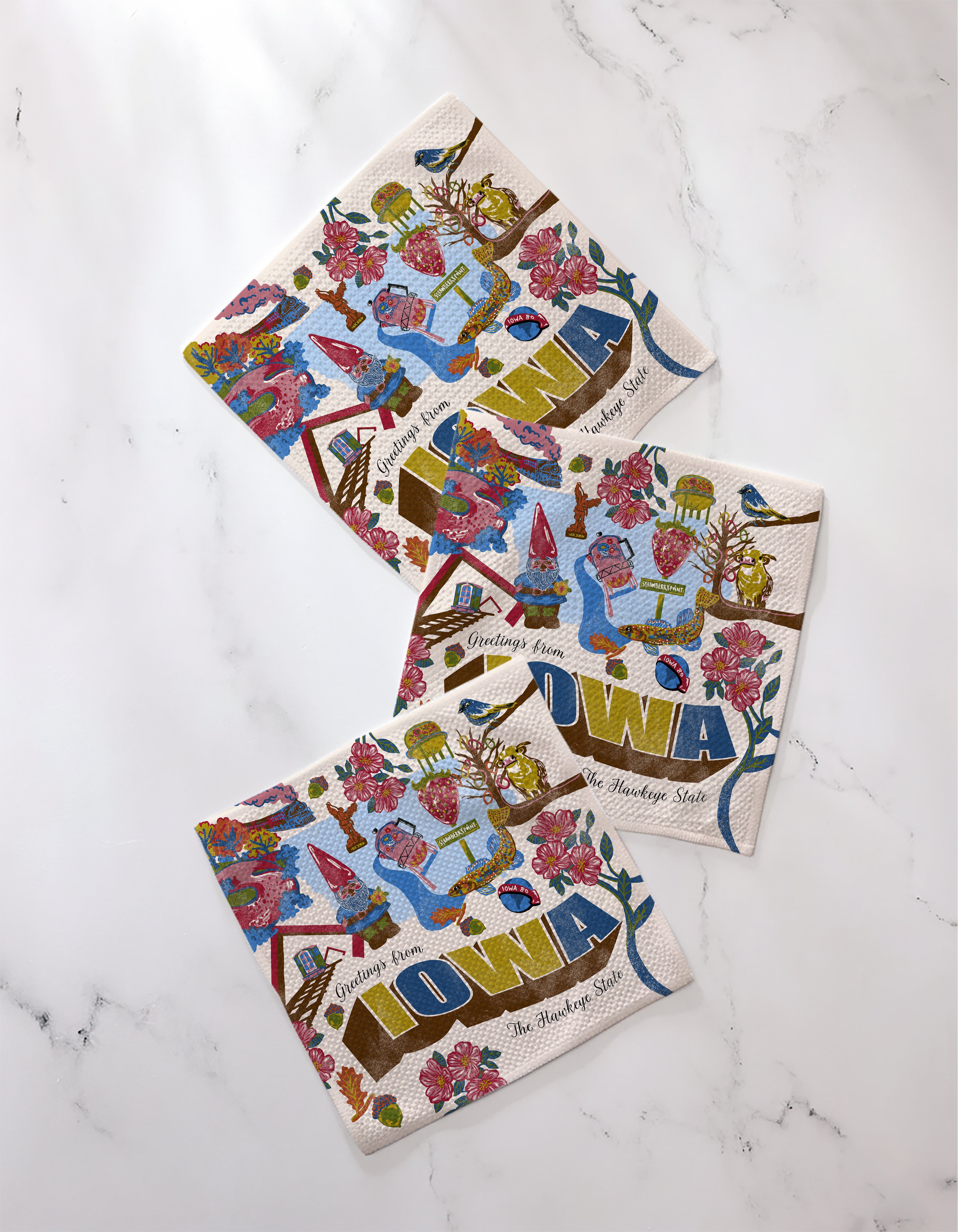 The Geometry "Greetings From Iowa" decorative paper napkins feature recycled materials and colorful Iowa-themed illustrations, including flowers, birds, barns, and the phrase “The Hawkeye State,” all displayed on a white marble surface.