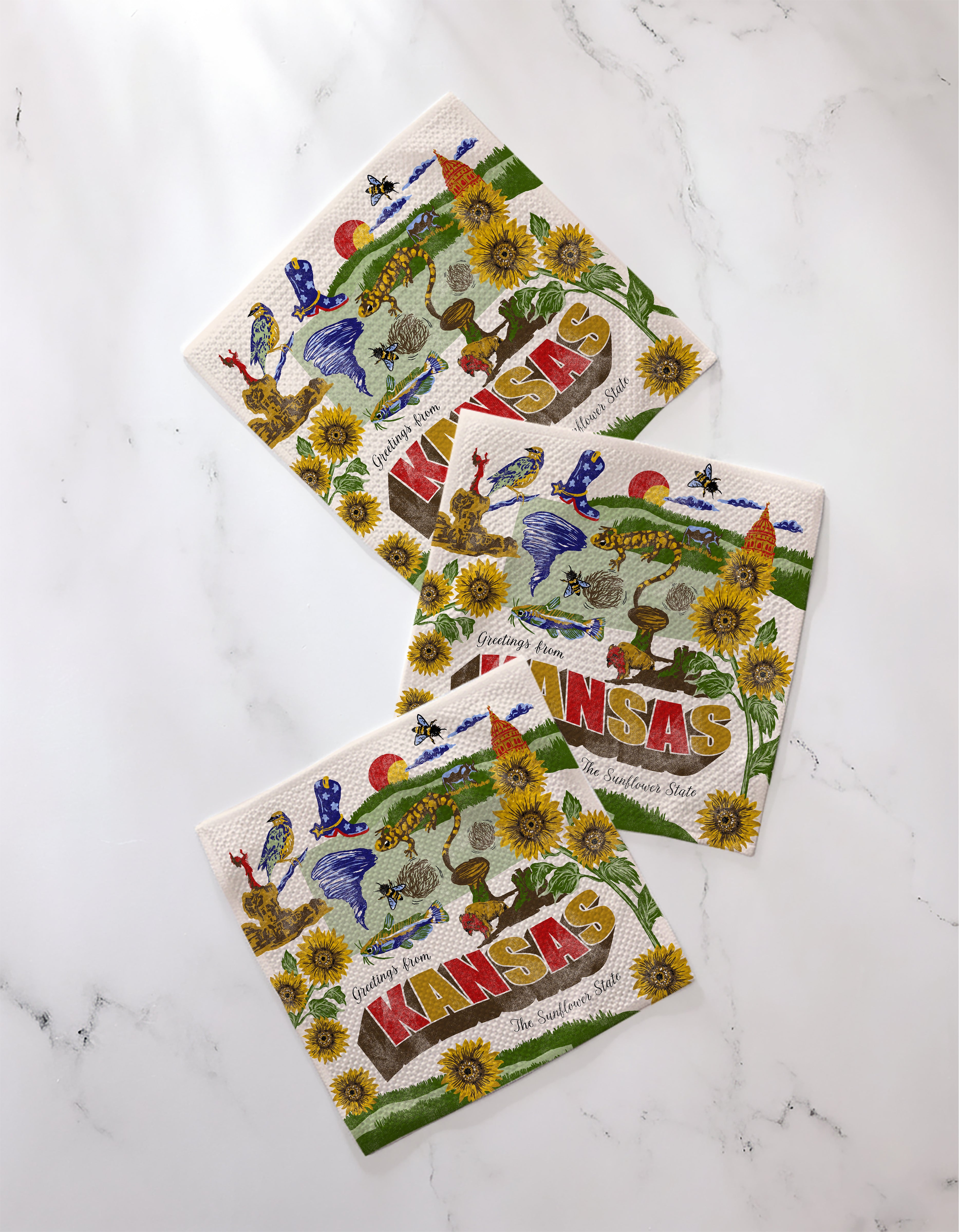 Three “Greetings From Kansas” paper napkins by Geometry, made from recycled materials, lie on a white marble surface. Each features vibrant sunflowers, birds, a red "KANSAS," and a state map outline.