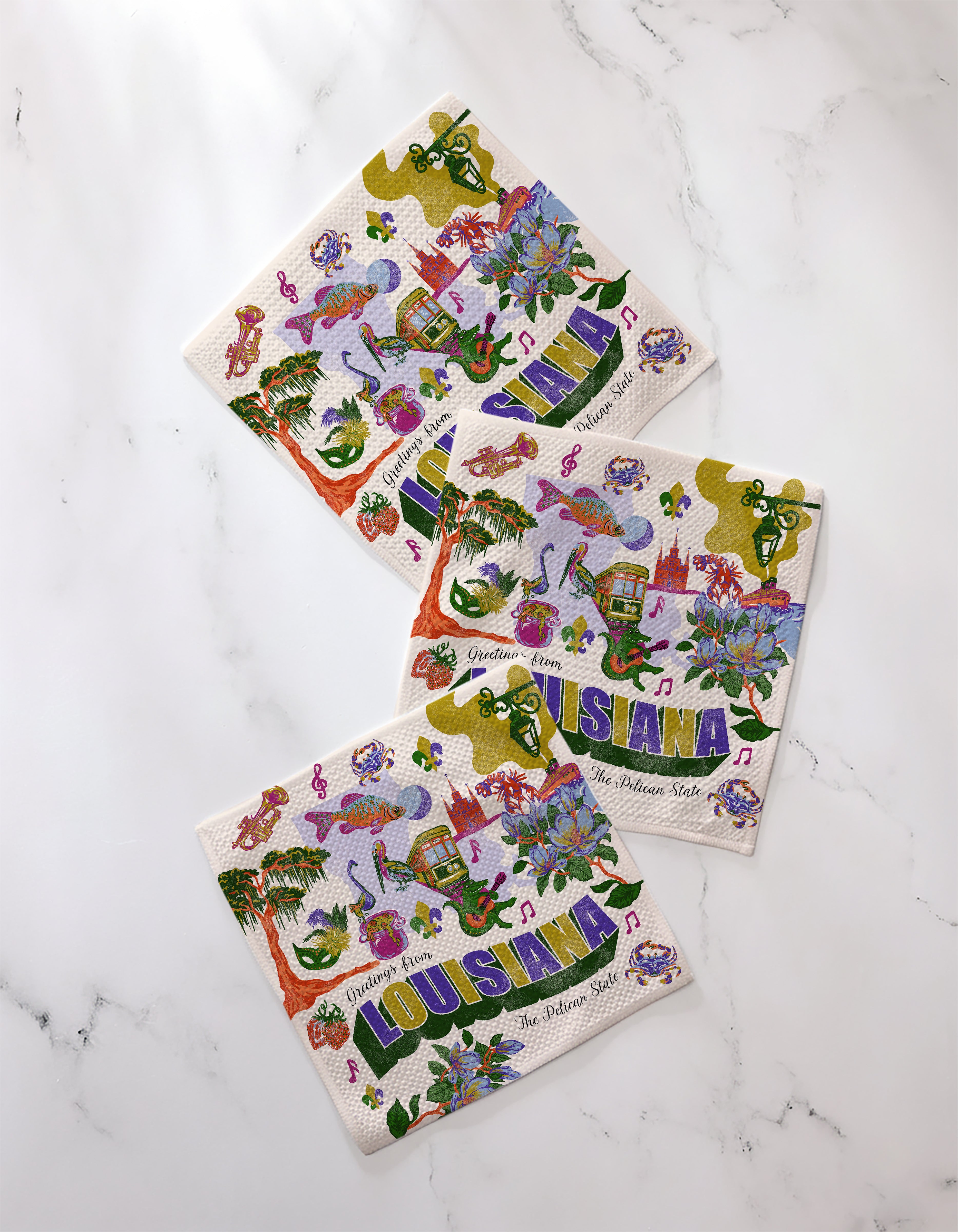Three vibrant Geometry dishcloths from the Greetings From Louisiana collection—featuring musical notes, alligators, and Mardi Gras masks—are arranged on white marble. Each recycled cloth displays "LOUISIANA" prominently.