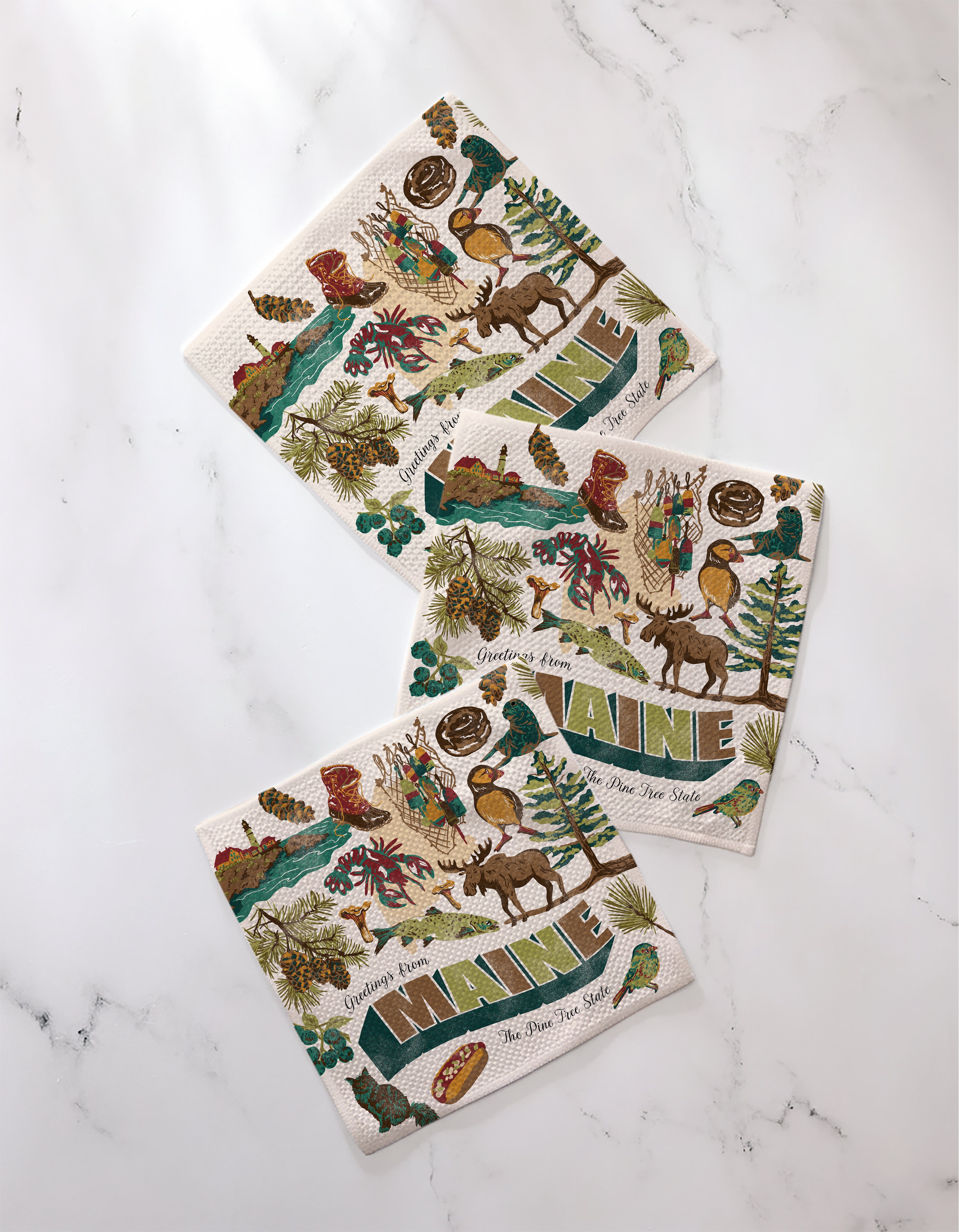 Three Geometry “Greetings From Maine” recycled kitchen towels, brightly illustrated with moose, trees, mountains, lobsters, and MAINE text, are arranged on white marble—perfect for any Maine-loving home.