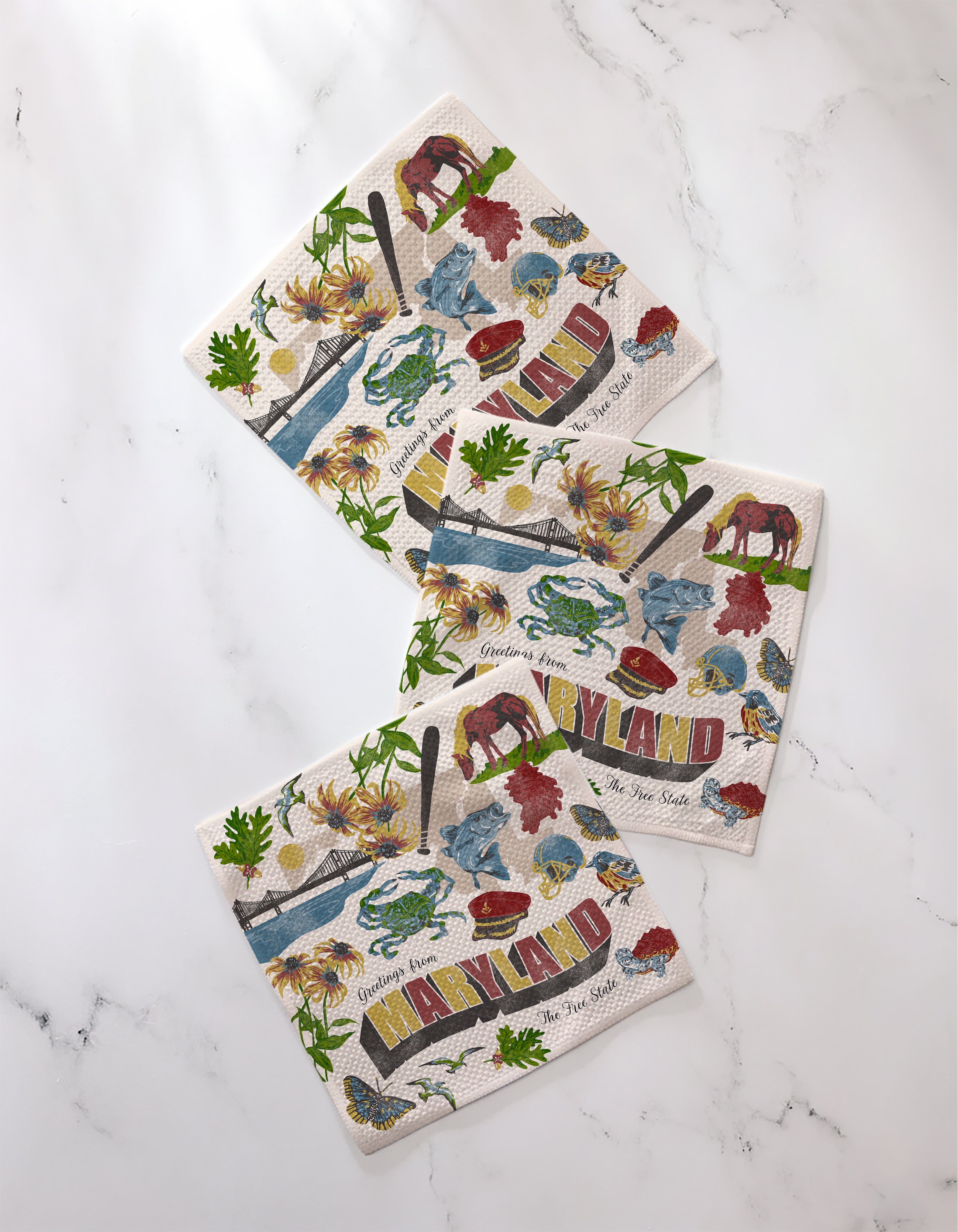 Three "Greetings From Maryland" napkins by Geometry, featuring colorful state symbols like blue crabs, a lighthouse, and a sailboat, are made from recycled materials and displayed on white marble.