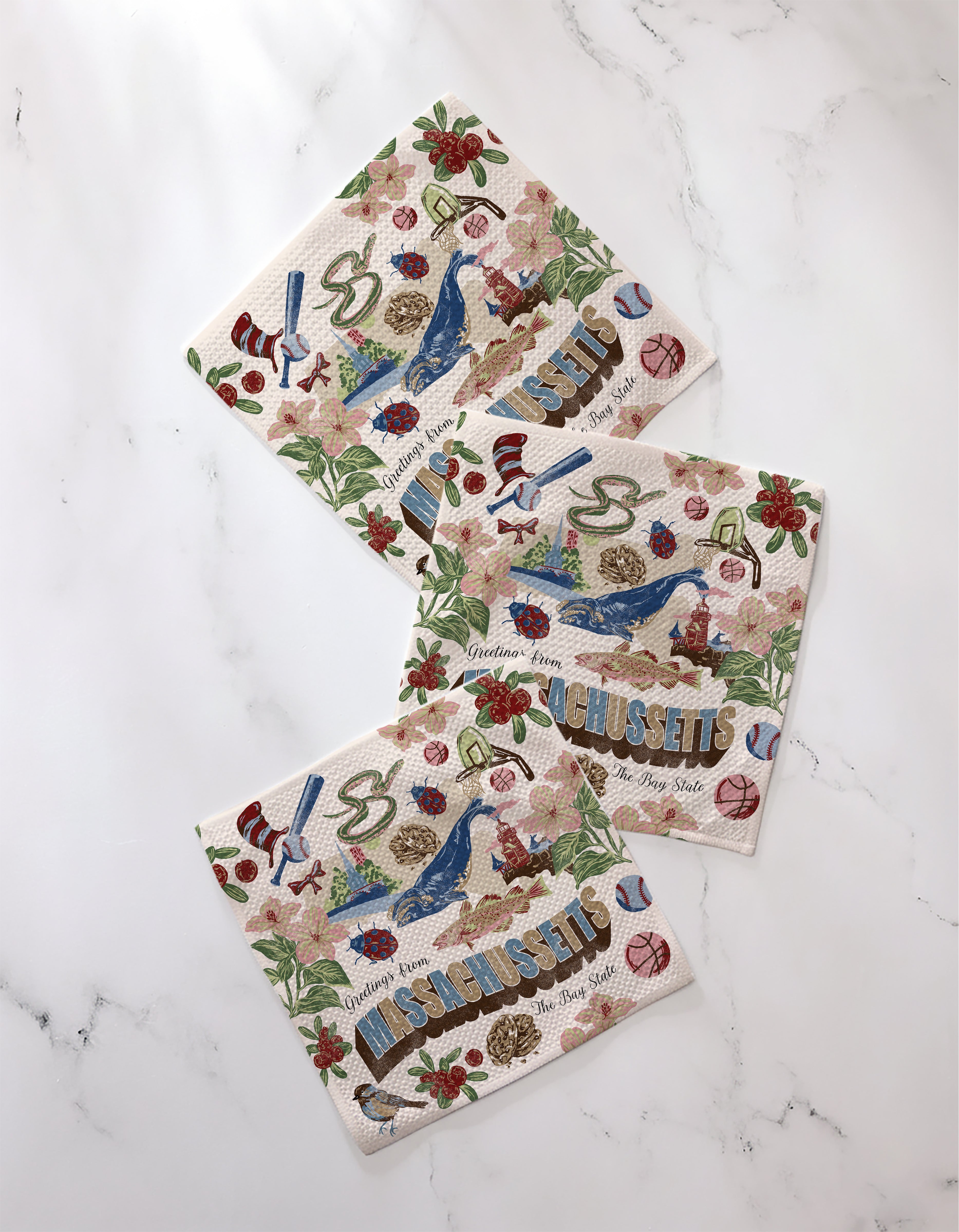 Three "Greetings From Massachusetts" paper napkins by Geometry, featuring state-inspired illustrations, are displayed on white marble. Pair with Geometry’s super-absorbent dishtowels for a charming kitchen accent.