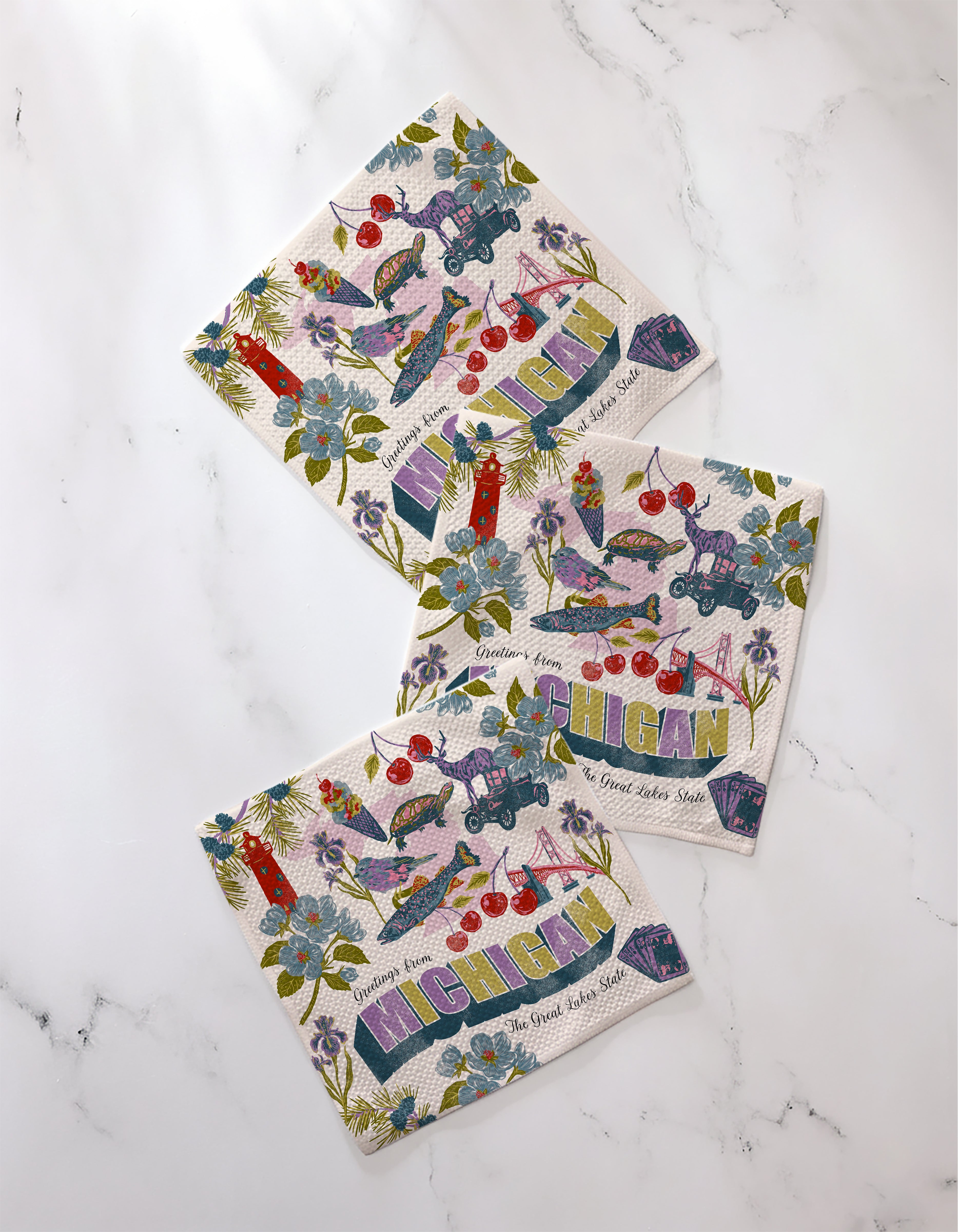 Three vibrant Geometry napkins from the "Greetings From Michigan" collection, featuring Michigan motifs like flowers, cherries, a car, and landmarks, are displayed on white marble. Made from recycled materials and labeled "Michigan, The Great Lakes State.