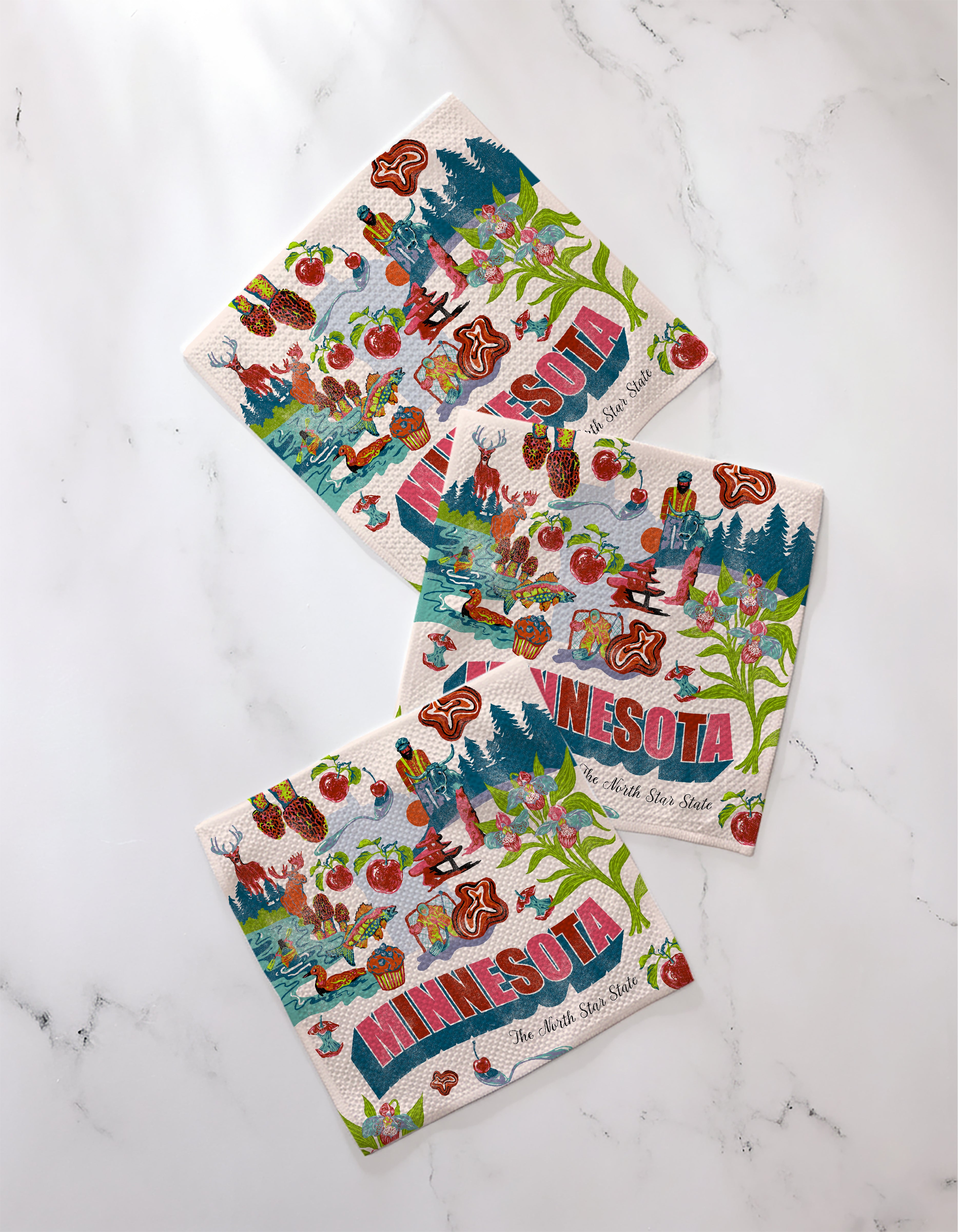 Three "Greetings From Minnesota" napkins by Geometry, made from recycled kitchen towels and decorated with colorful Minnesota lakes, trees, animals, and symbols, are shown on white marble with “MINNESOTA” displayed.