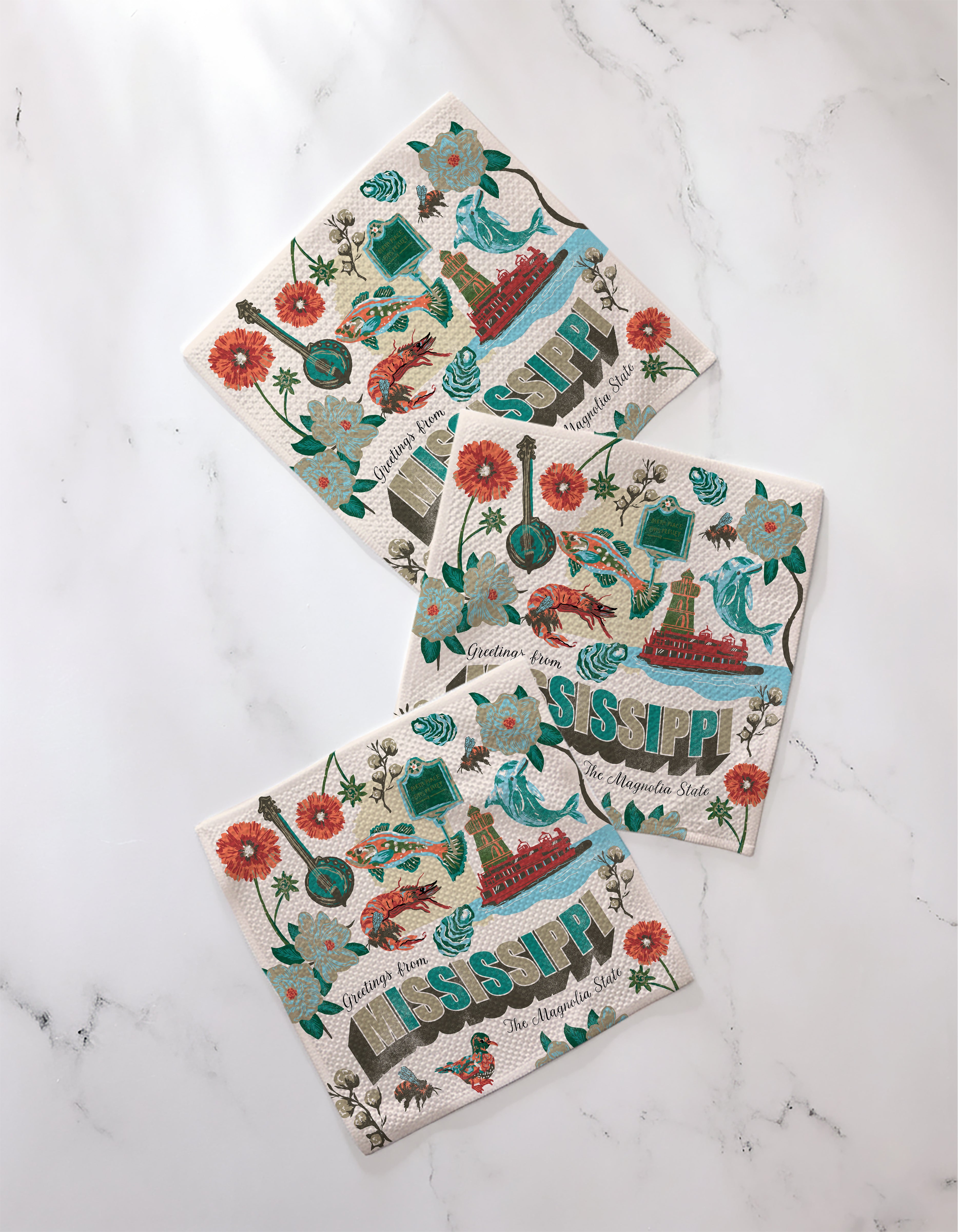 The Geometry "Greetings From Mississippi" set includes three decorative napkins made from recycled materials, each featuring vibrant illustrations of flowers, birds, musical instruments, and other Mississippi-inspired elements.