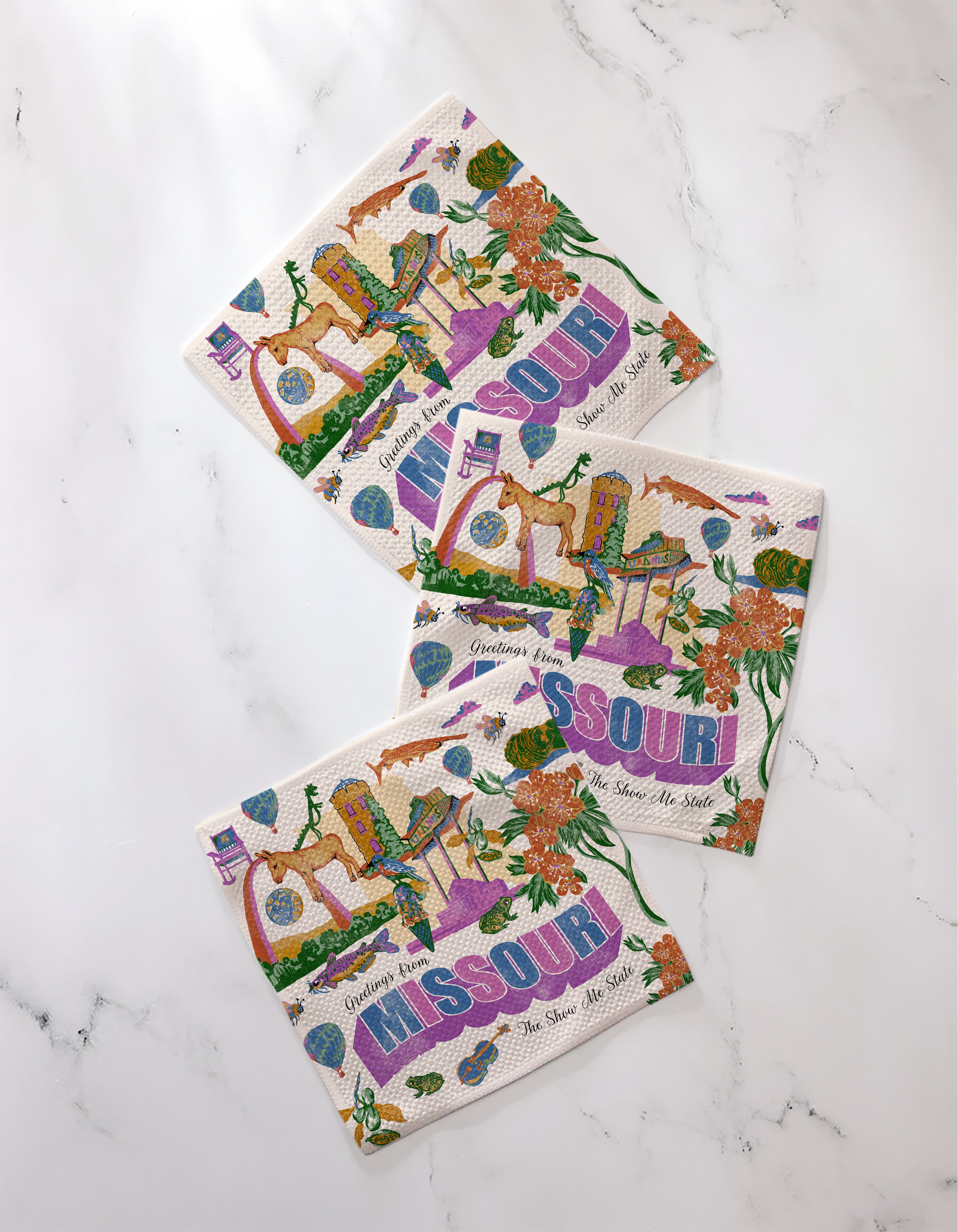 Three vibrant “Greetings From Missouri” paper napkins by Geometry, featuring the state’s flowers, birds, and river, are arranged on white marble—perfectly complementing any recycled-material dishcloth set.
