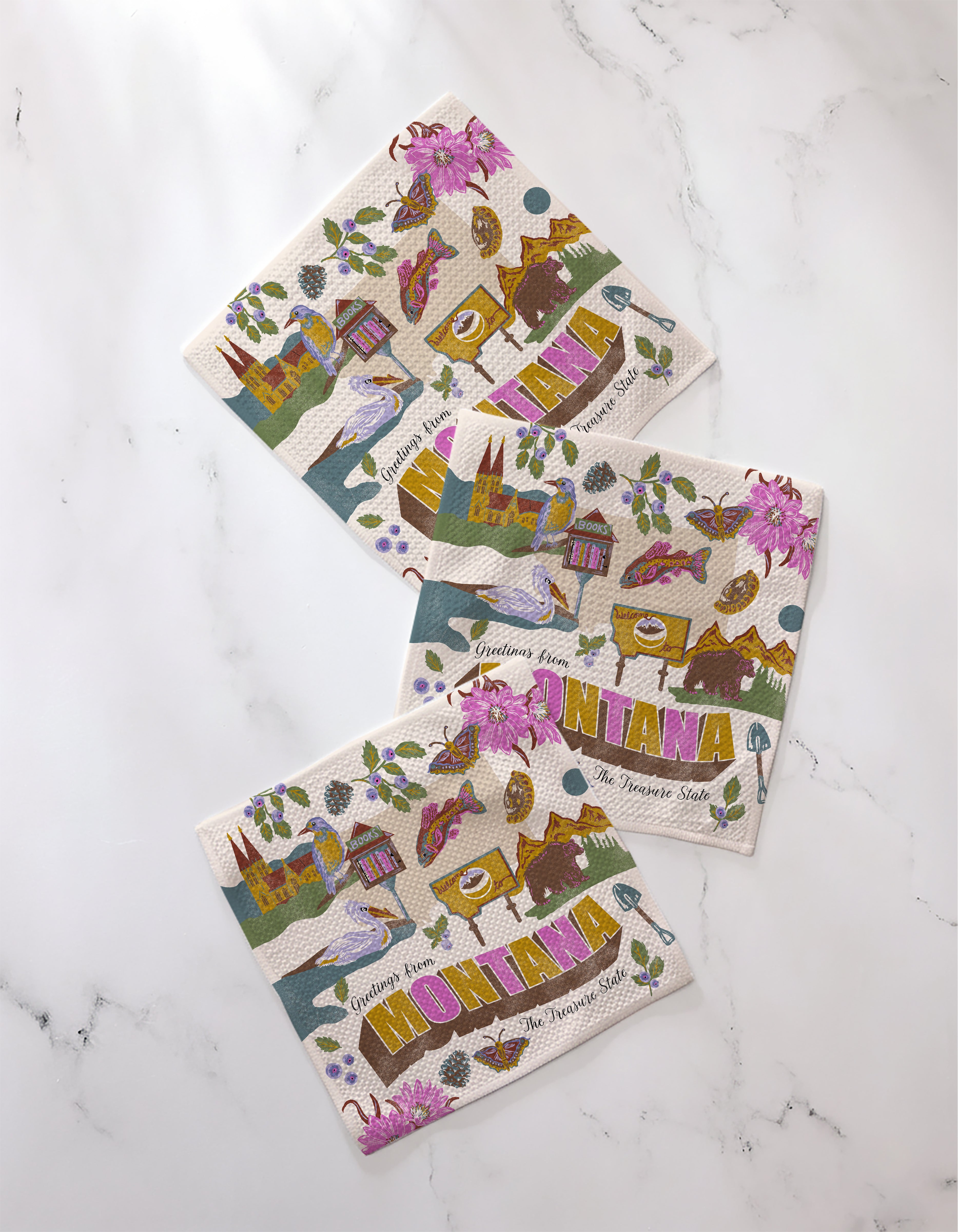 Three colorful "Greetings From Montana" paper napkins by Geometry are displayed on white marble. Pair them with a matching absorbent dishcloth set for a charming, practical table setting.