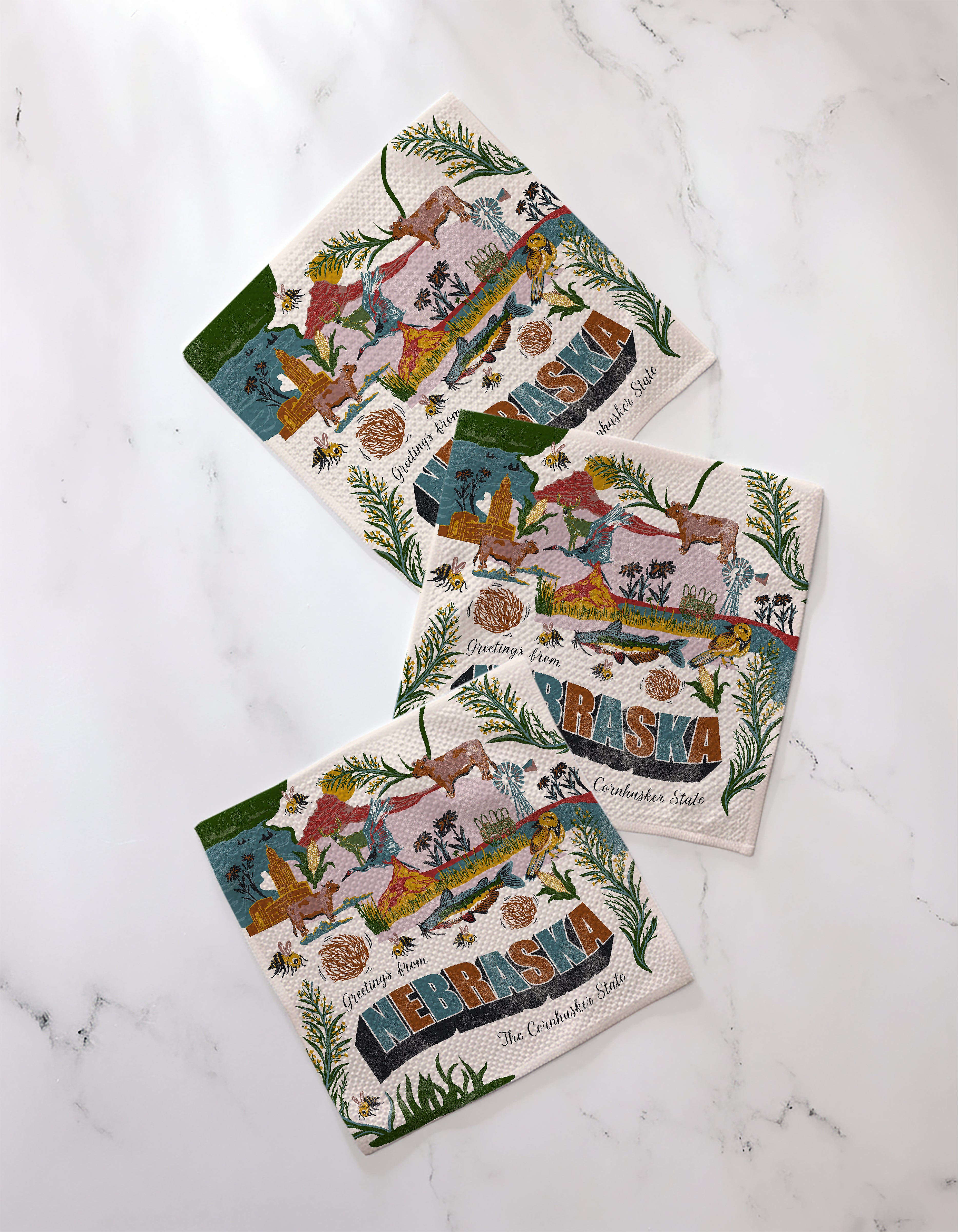 Three vibrant, super-absorbent Geometry dishtowels from the "Greetings From Nebraska" collection—made with recycled materials and featuring Nebraska-inspired illustrations and bold NEBRASKA text—are arranged on a white marble surface.