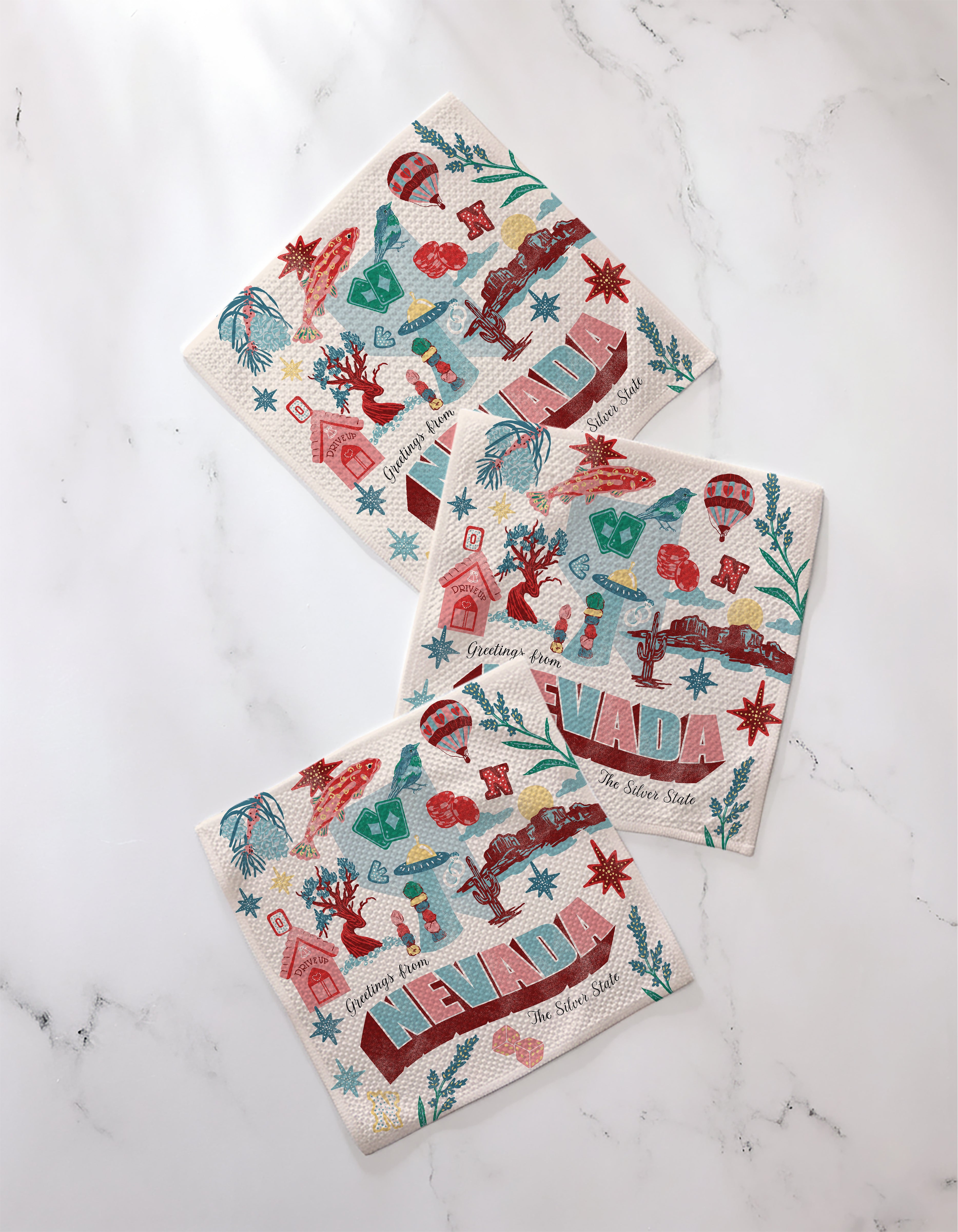 The Geometry "Greetings From Nevada" set includes three decorative paper napkins featuring colorful Nevada-themed illustrations—cacti, cowboy boots, and vibrant NEVADA text—beautifully displayed on a white marble surface.