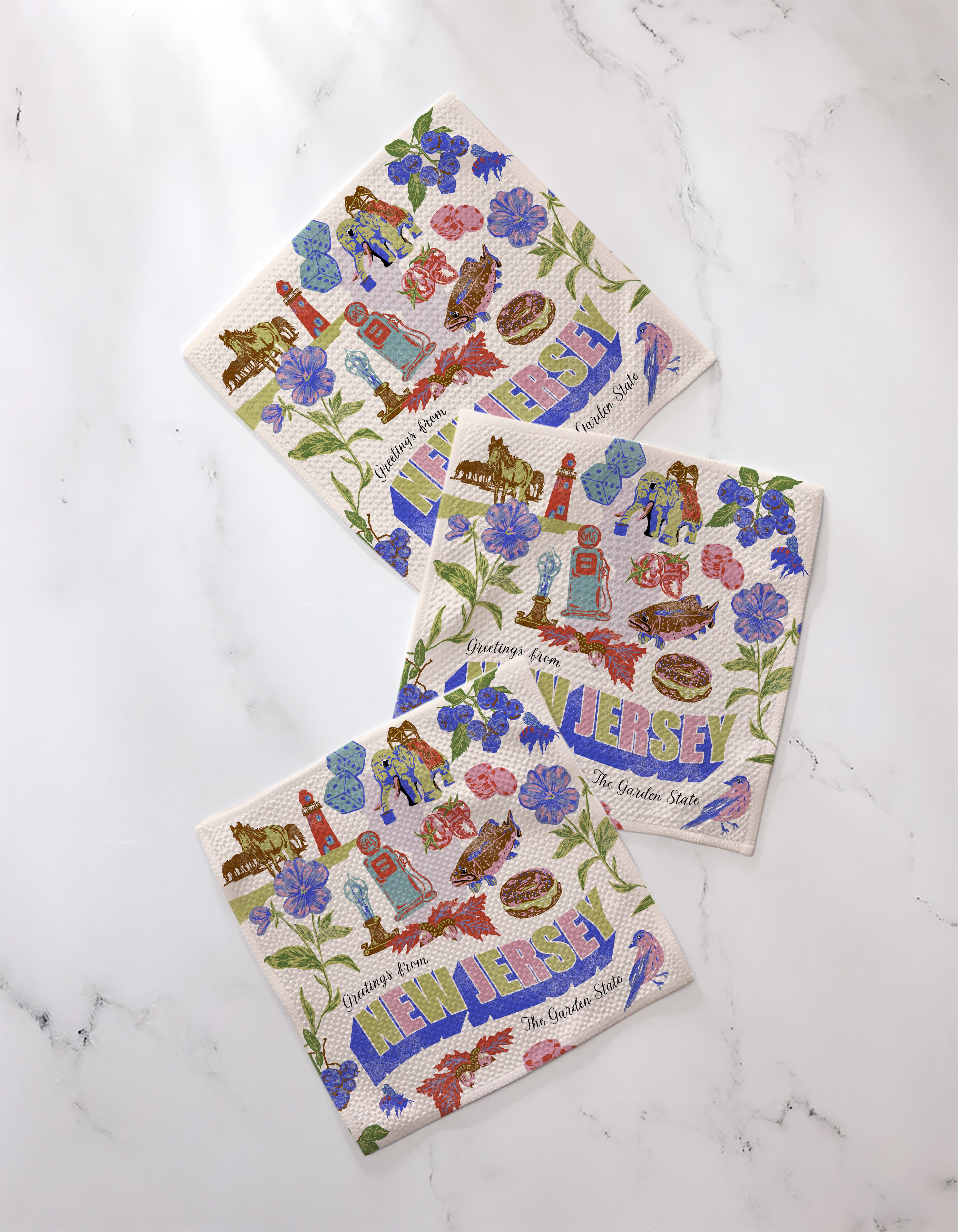 Three decorative recycled dishcloths from Geometry’s "Greetings From New Jersey" collection rest on white marble, featuring colorful illustrations of NJ symbols—flowers, horses, lighthouses—and the words “Greetings from New Jersey, The Garden State.”.