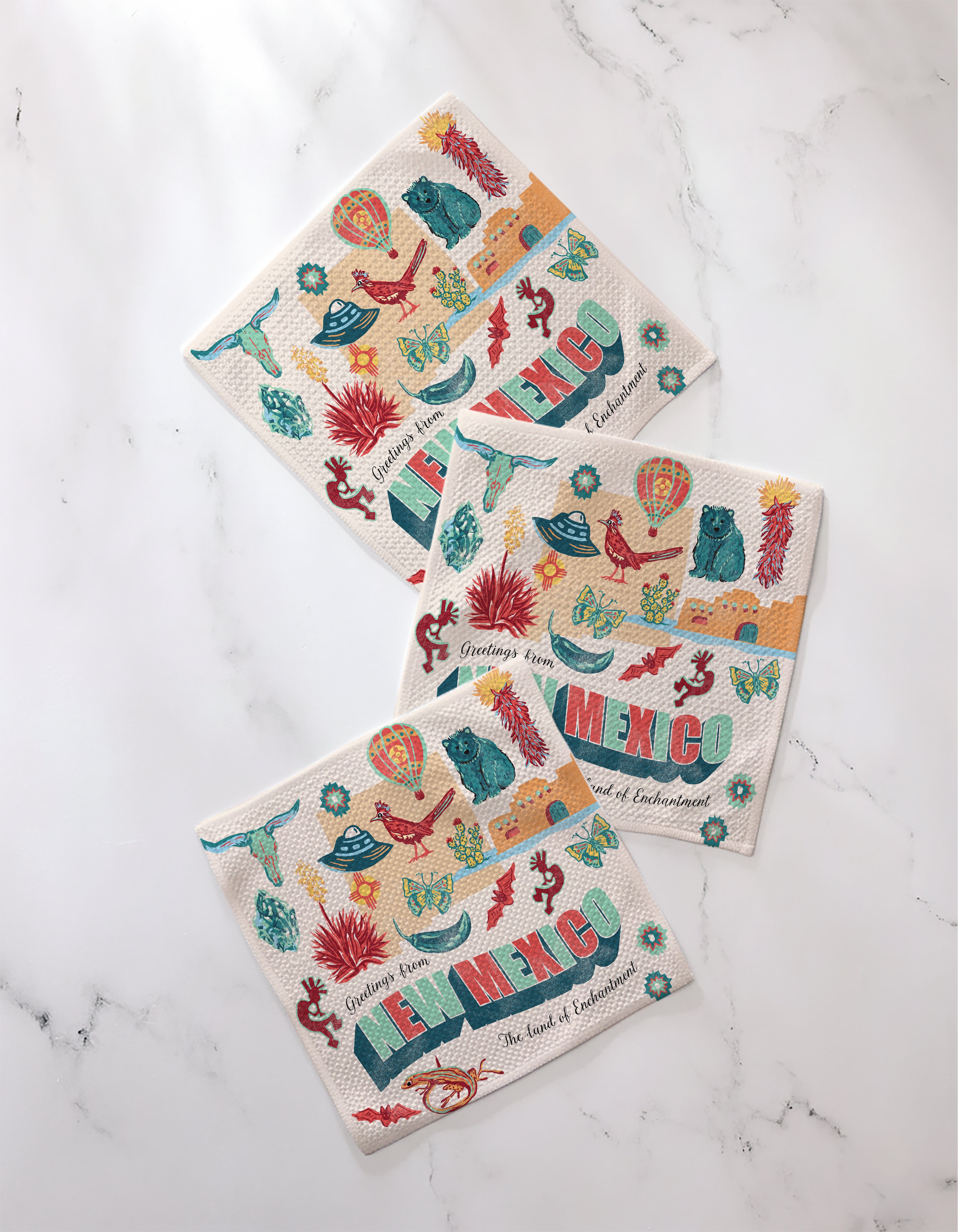 Geometry's Greetings From New Mexico decorative paper napkins feature cacti, hot air balloons, chili peppers, and lizards, with colorful illustrations and text. Eco-friendly design made from recycled materials on a white marble surface.