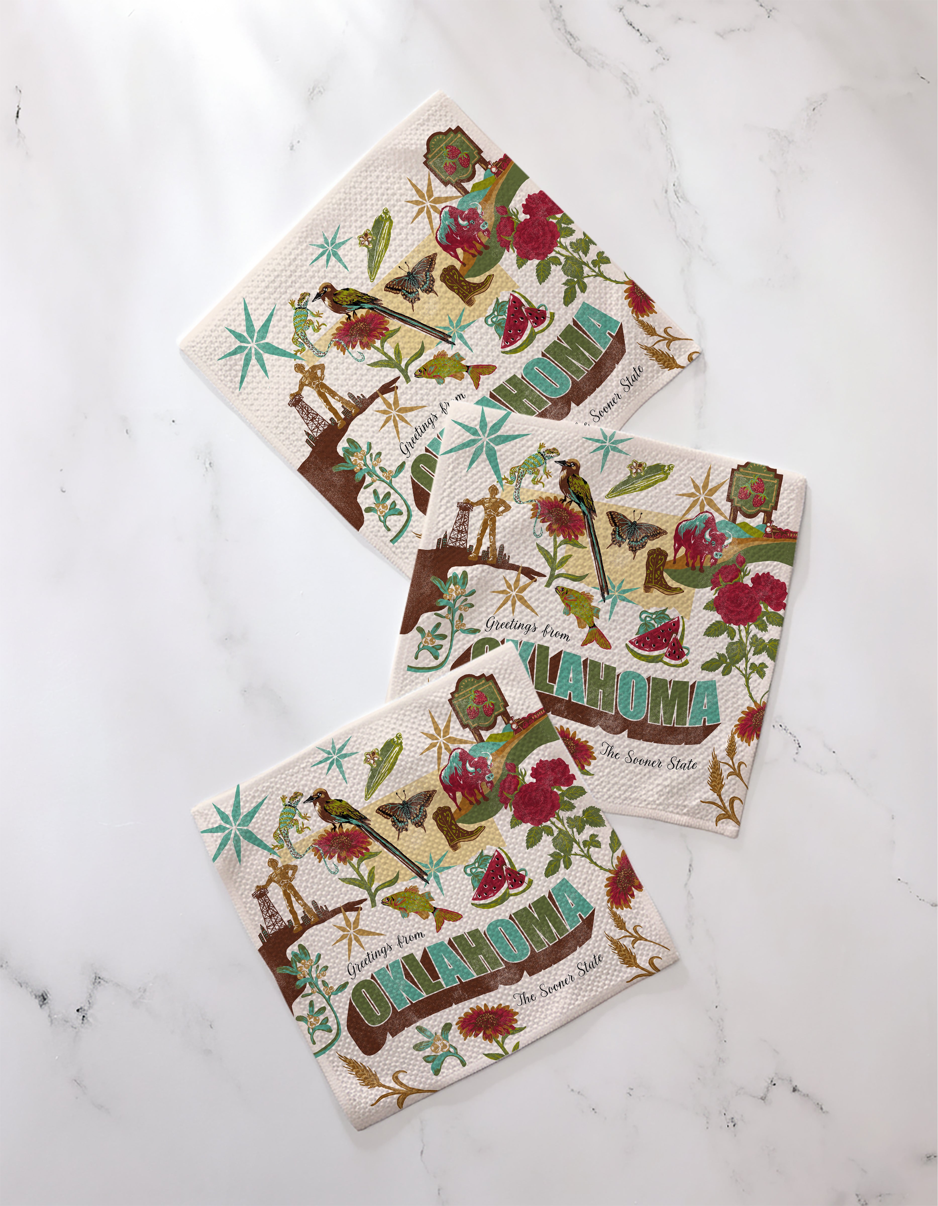 Three super-absorbent Geometry dishtowels from the "Greetings From Oklahoma" collection, featuring colorful state-themed illustrations, are neatly displayed on a white marble surface.