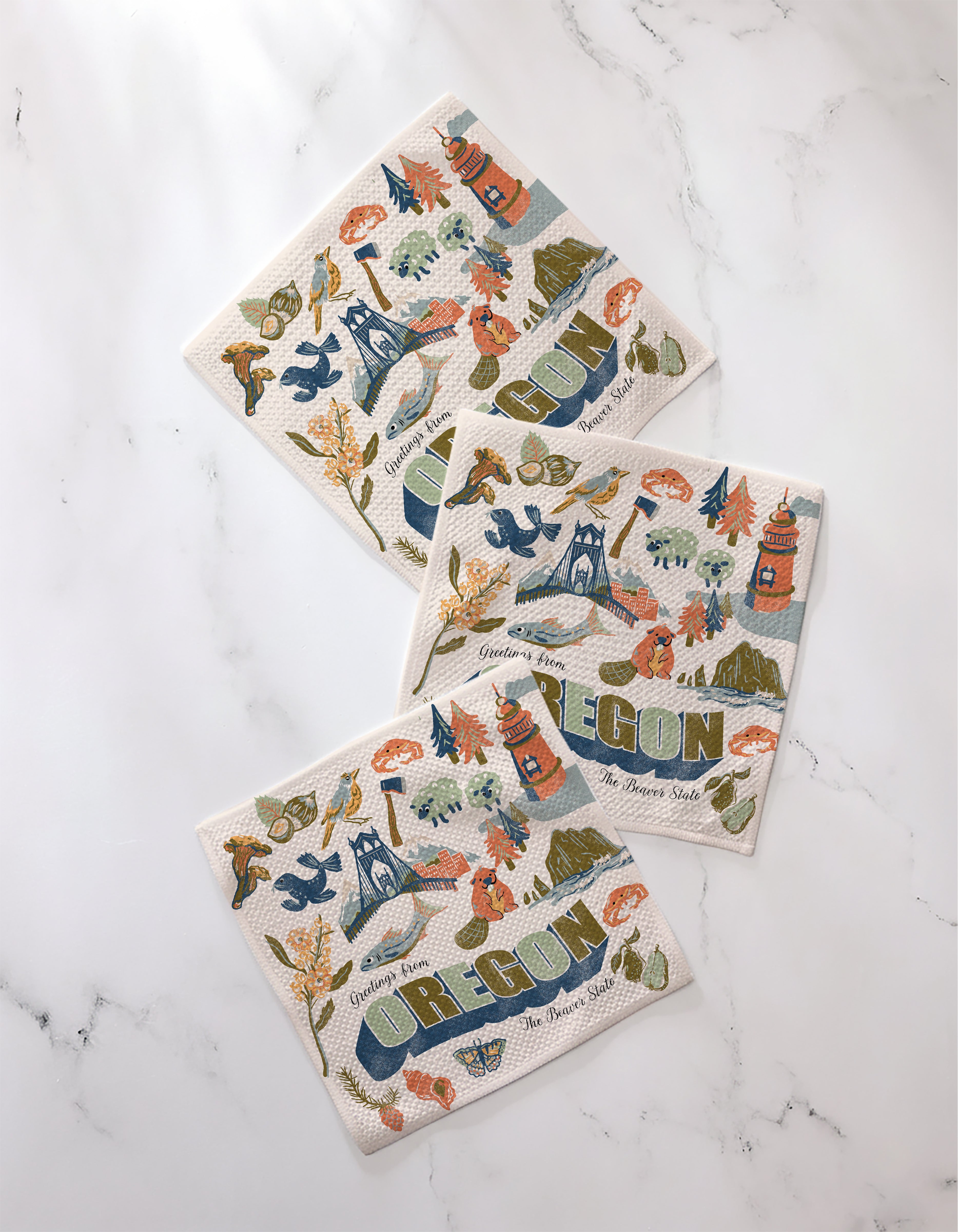 Three colorful, super-absorbent Geometry "Greetings From Oregon" dishtowels on a marble surface, each featuring OREGON and vibrant illustrations of state icons like trees, mountains, lighthouses, crabs, mushrooms, and flowers.
