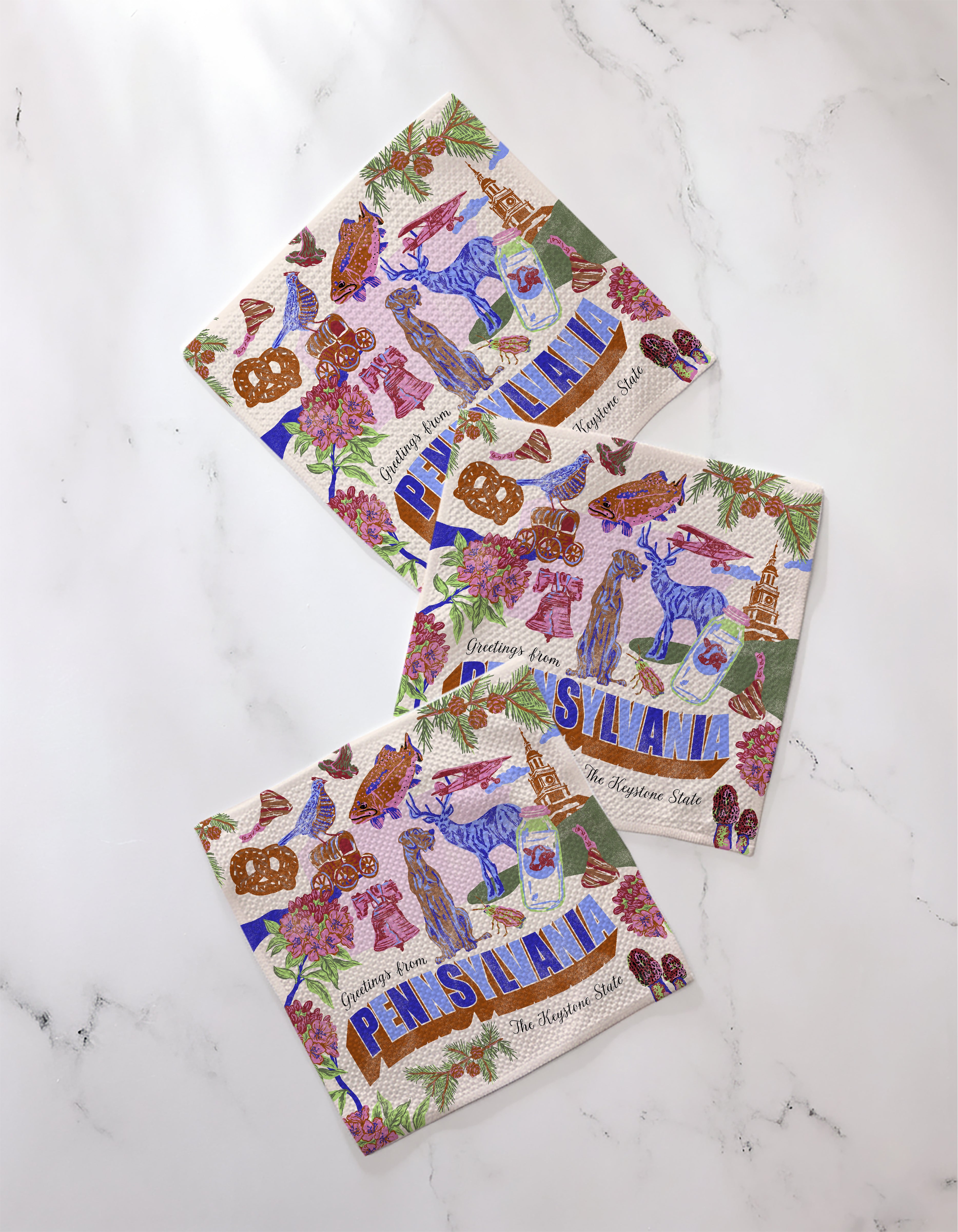 Three eco-friendly Geometry napkins, titled "Greetings From Pennsylvania," display illustrated state icons like pretzels and deer. The decorative set is arranged on a white marble surface.