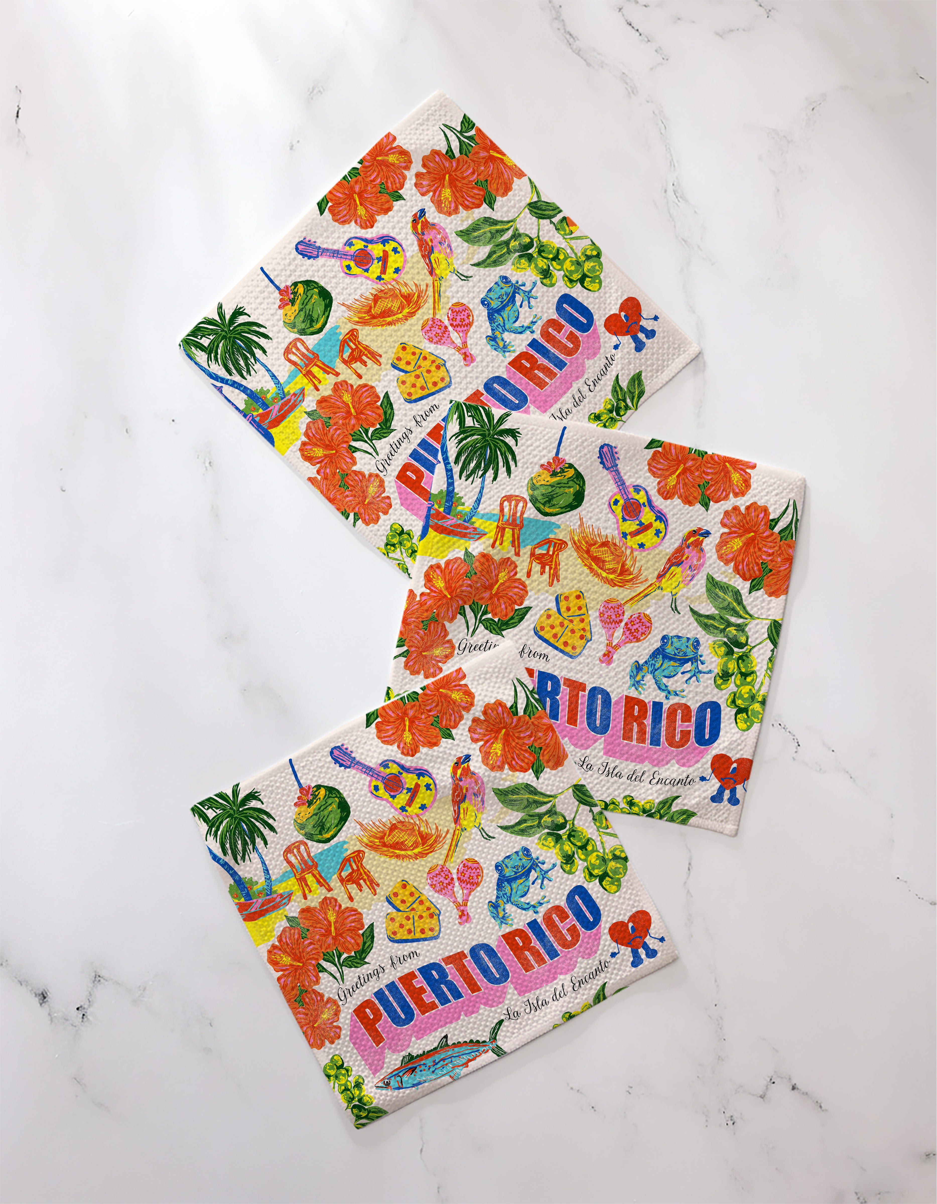 Three vibrant, eco-friendly Geometry "Greetings From Puerto Rico" kitchen towels rest on white marble, each featuring tropical flowers, fruits, palm trees, and “Puerto Rico” in bold blue letters.