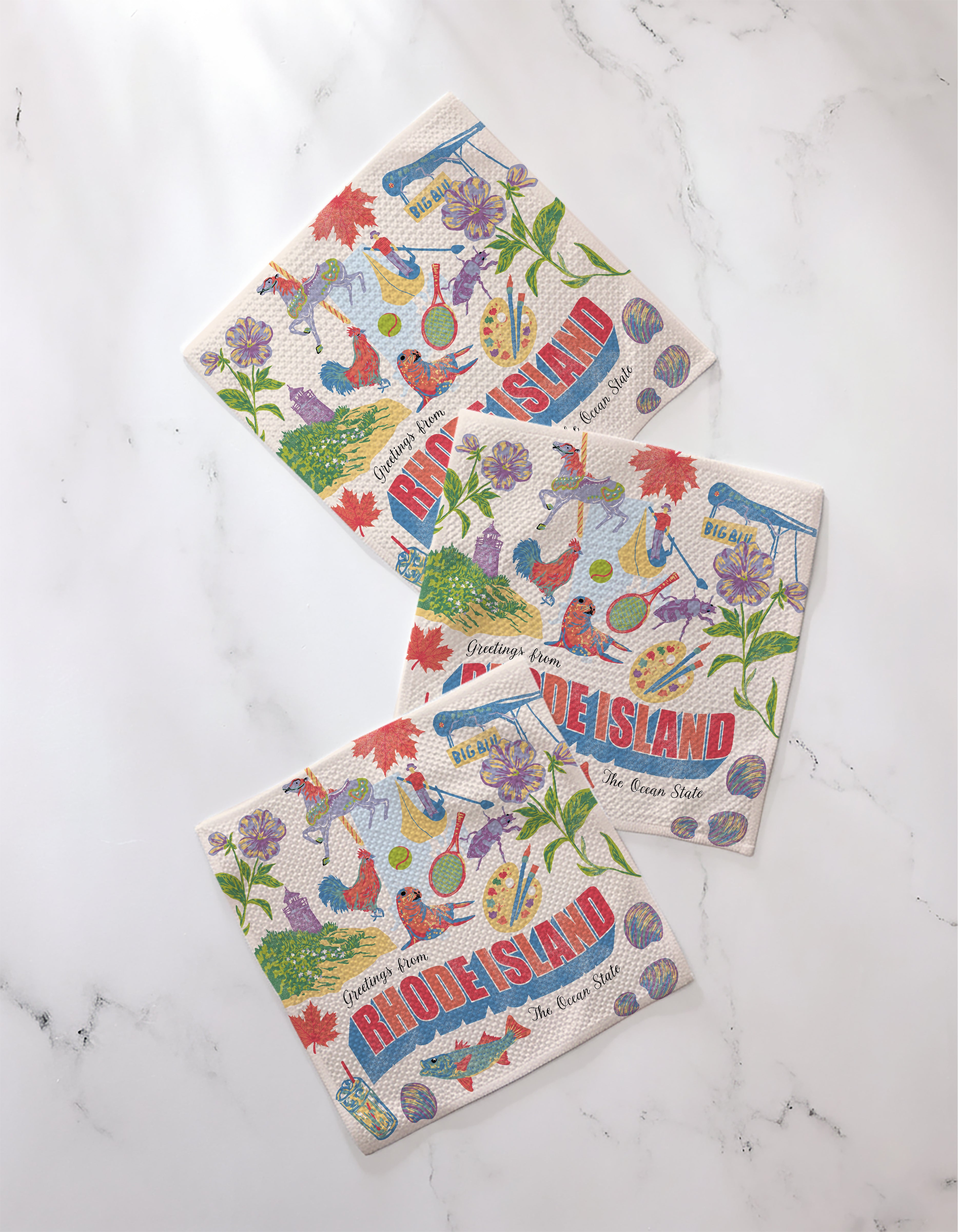 Three decorative, super-absorbent Geometry "Greetings From Rhode Island" recycled dishcloths featuring flowers, musical instruments, seafood, and state symbols are arranged on a white marble surface.