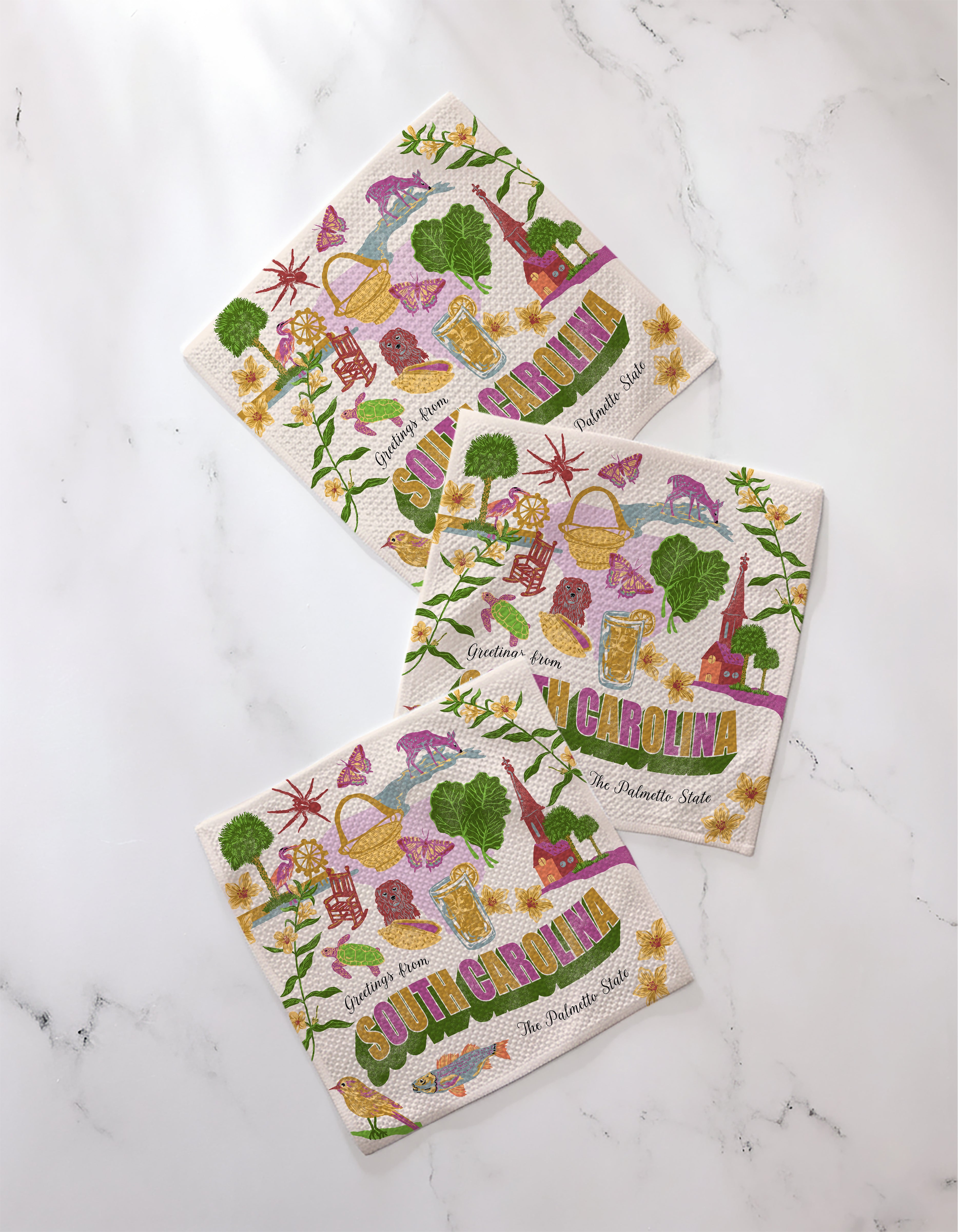 Three colorful Geometry “Greetings From South Carolina” dishcloths, featuring flowers, animals, landmarks, and the words “South Carolina” and “The Palmetto State,” are arranged on a white marble surface.