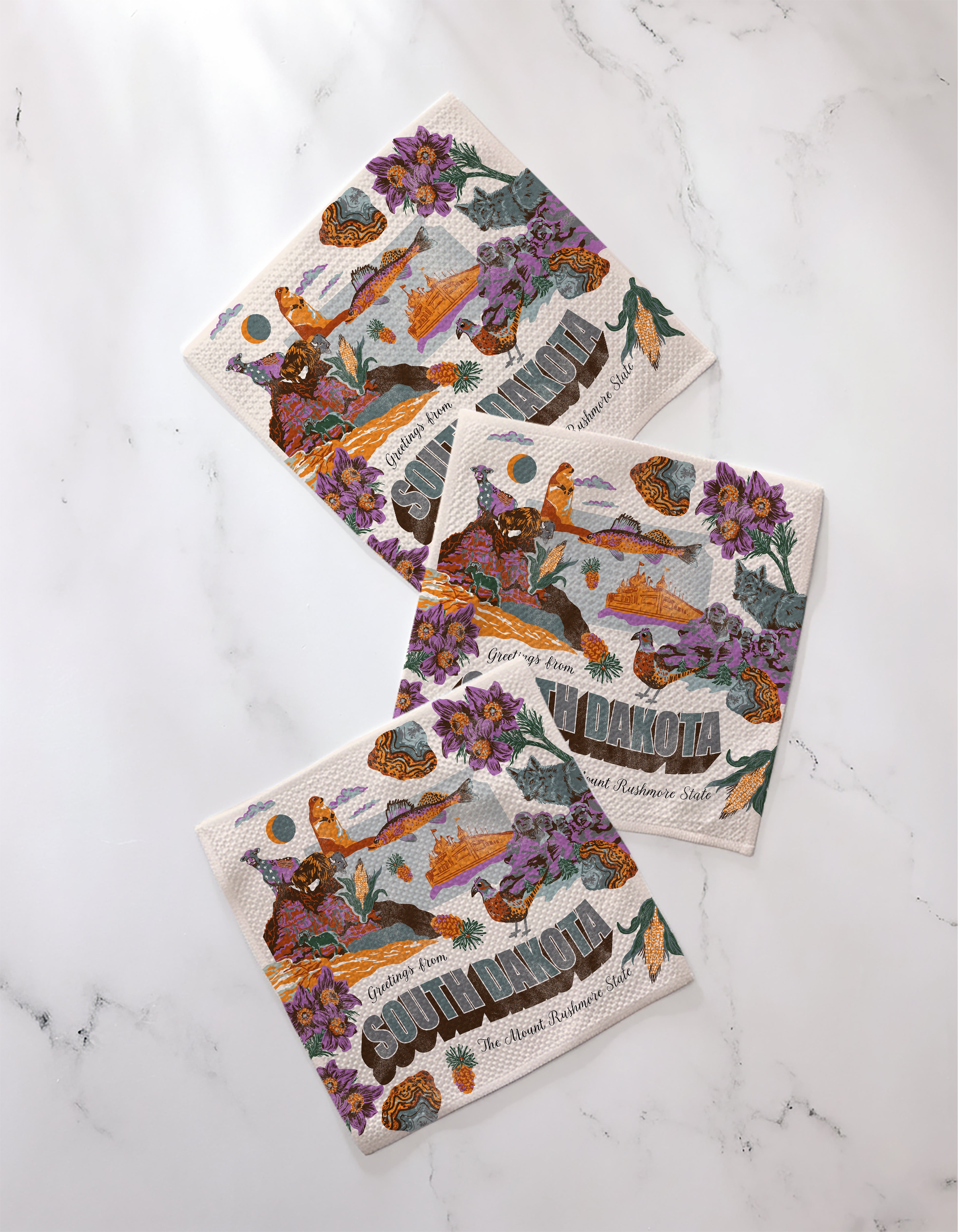 Three "Greetings From South Dakota" paper napkins by Geometry, featuring purple flowers, orange birds, and landscape art, are displayed on white marble—perfectly paired with a set of super-absorbent dishtowels for any kitchen.