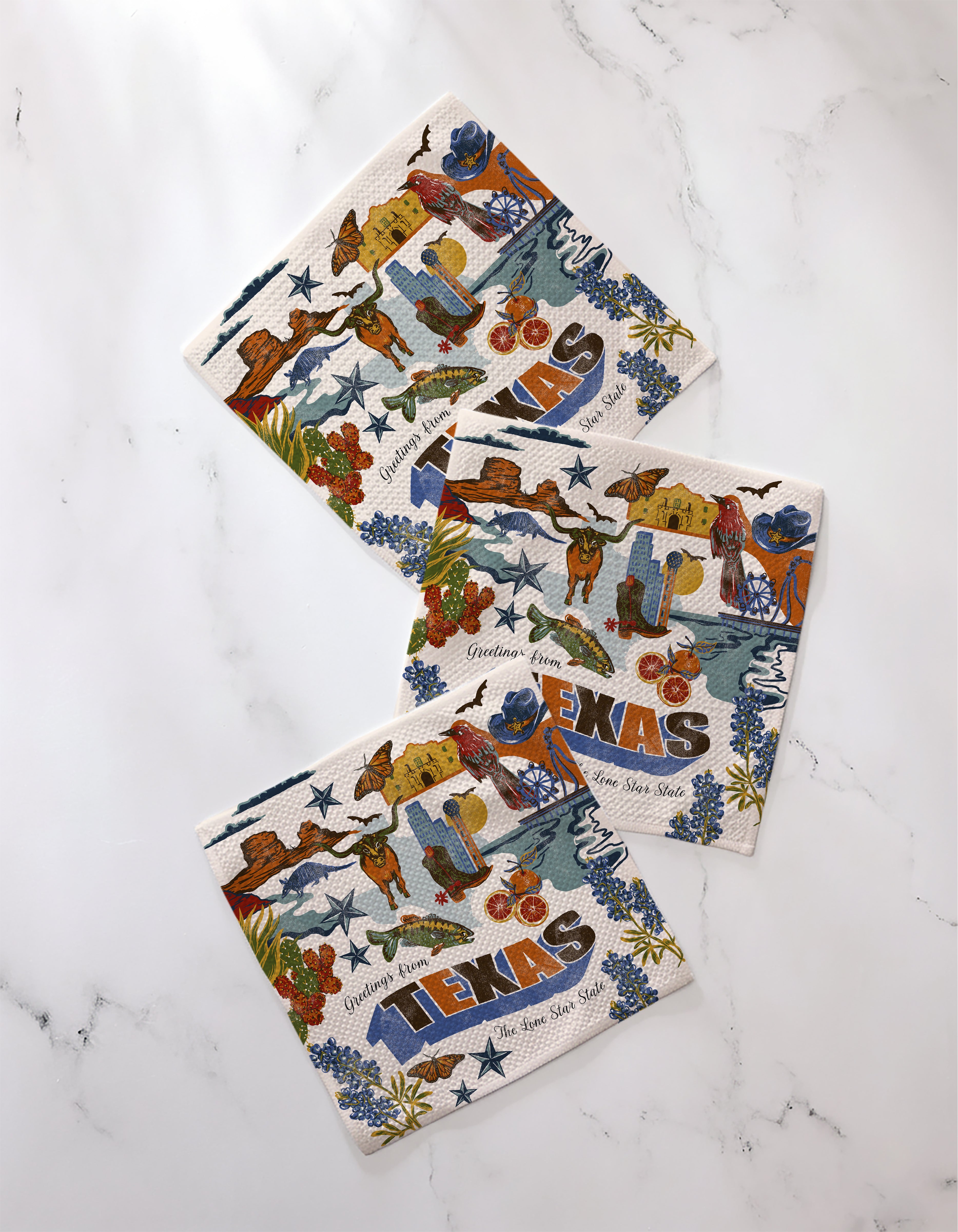 The Geometry "Greetings From Texas" set includes three recycled decorative napkins featuring colorful Texas icons—a cowboy, cactus, armadillo, bluebonnets, and “TEXAS”—displayed on a white marble surface.